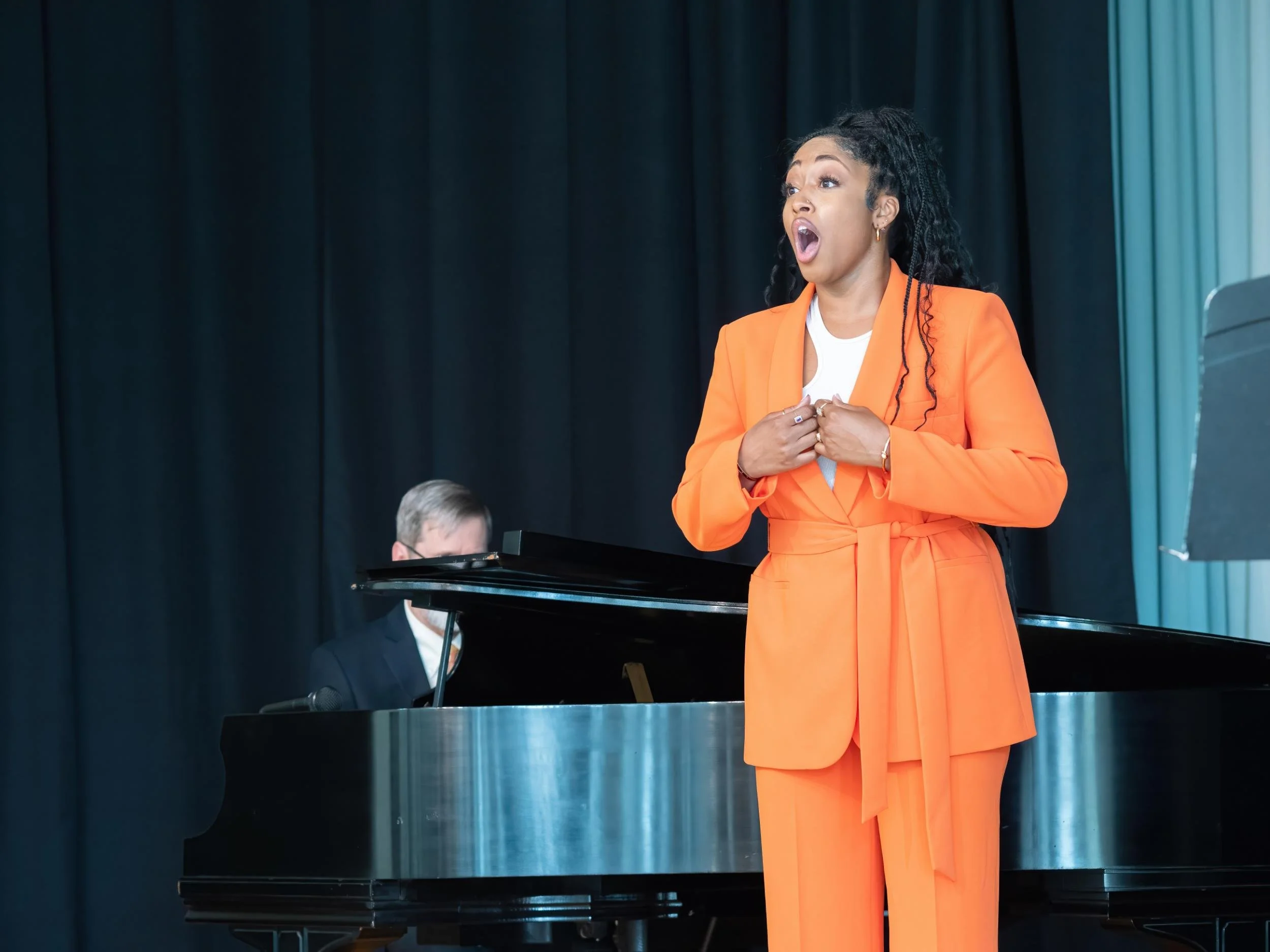 A woman in an orange suit singing passionately on stage while a man plays the piano in the background.