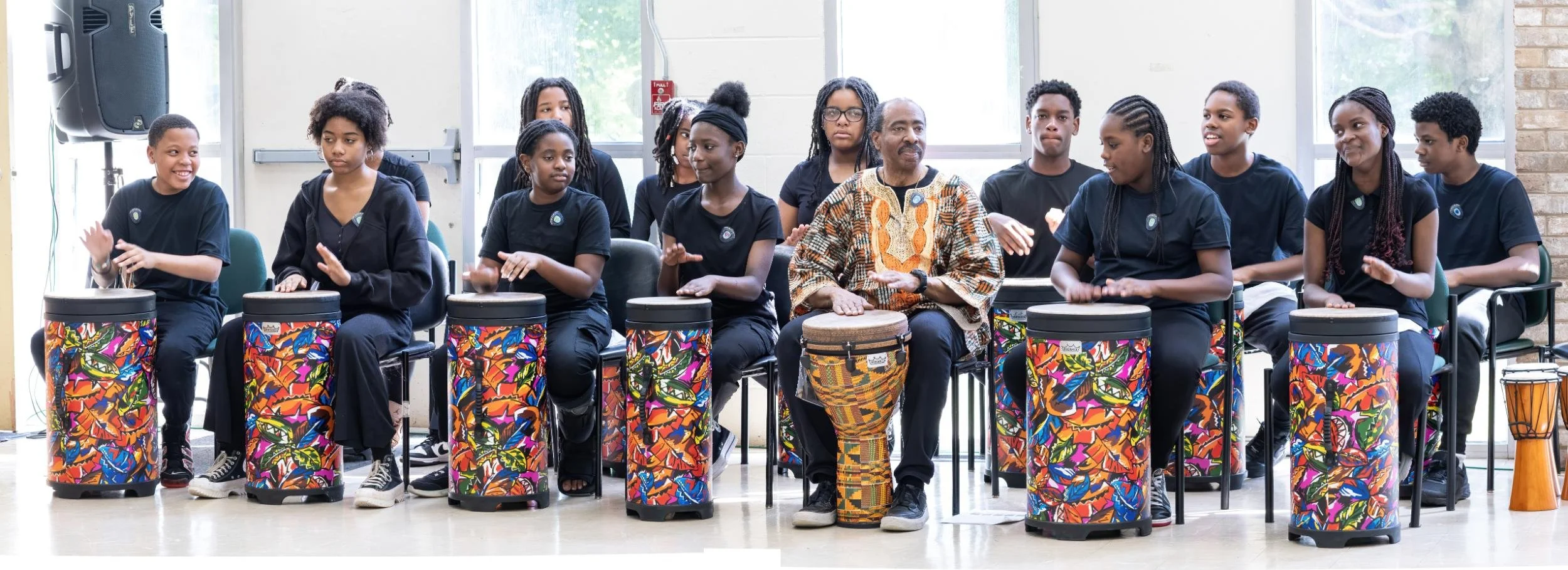 A diverse group of people, mainly young women, sitting in a row, each with a colorful drum in front of them. They are in a well-lit room with large windows and are engaged in music or drumming activity, with some clapping or playing their drums.