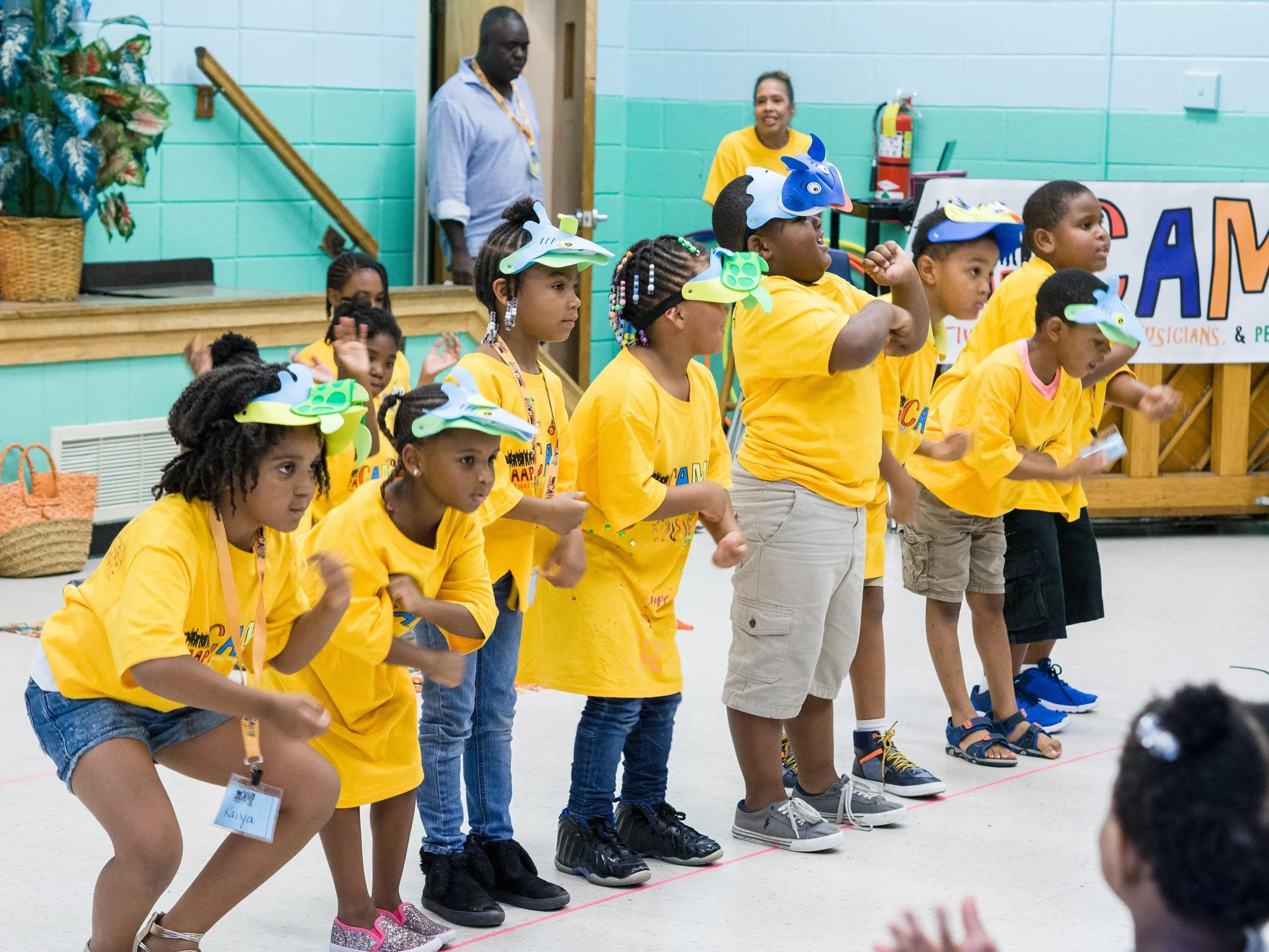 Children wearing yellow T-shirts and colorful bird masks perform on stage during a school or community event, with adults in the background and a banner reading 'CAM'.