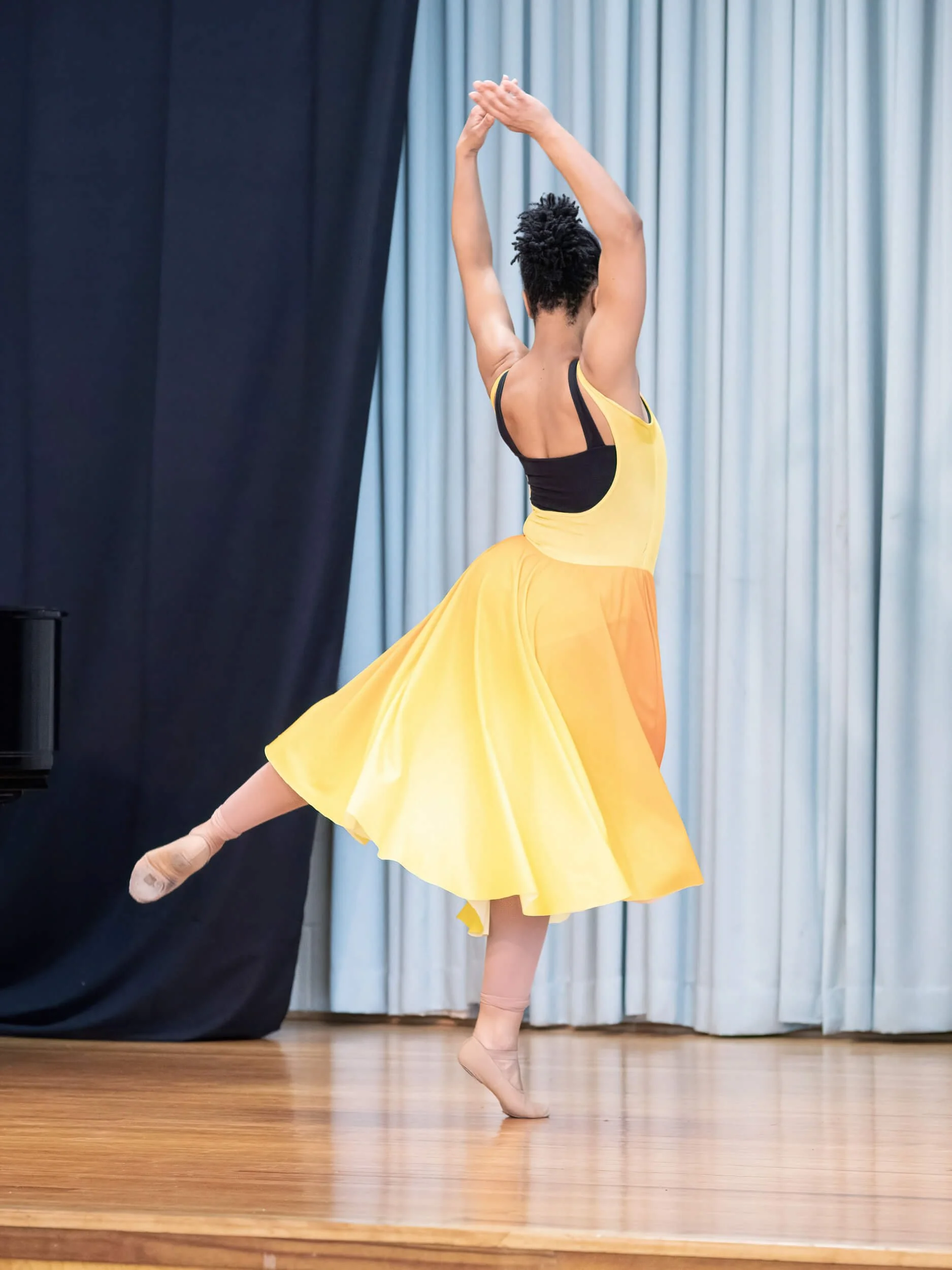 A dancer in a yellow dress and black top practicing ballet on a stage with blue curtains in the background.