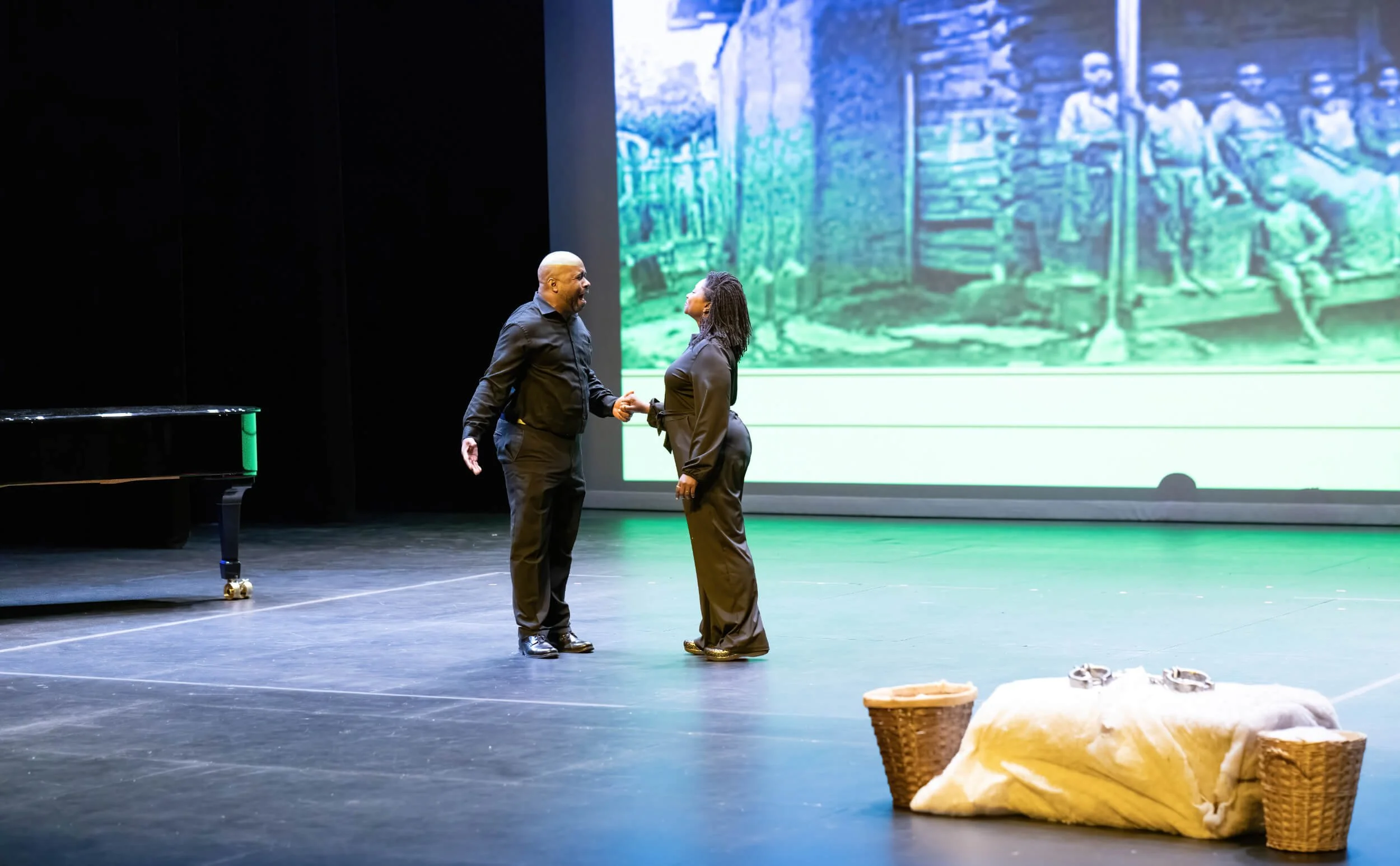 Two actors on stage holding hands, engaged in a dramatic scene, with a large projector screen showing a black and white photo of children playing in the background. There is a basket, a white blanket, and a pair of shoes on the stage.