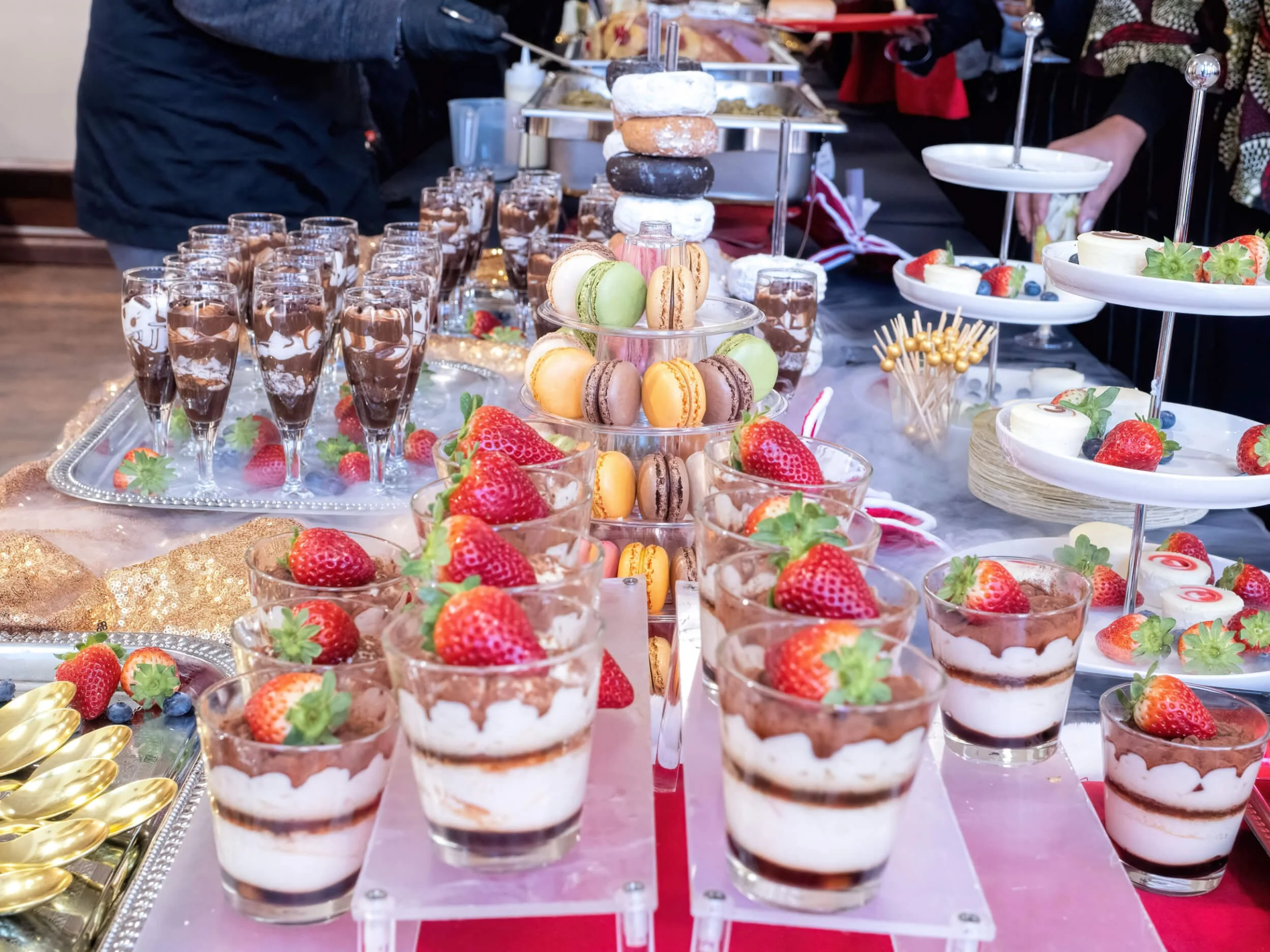 A dessert table with various sweet treats including strawberry parfaits topped with strawberries, layered macarons, chocolate mousse in glasses, and assorted fruits, arranged on multiple tiers and trays at a festive event.