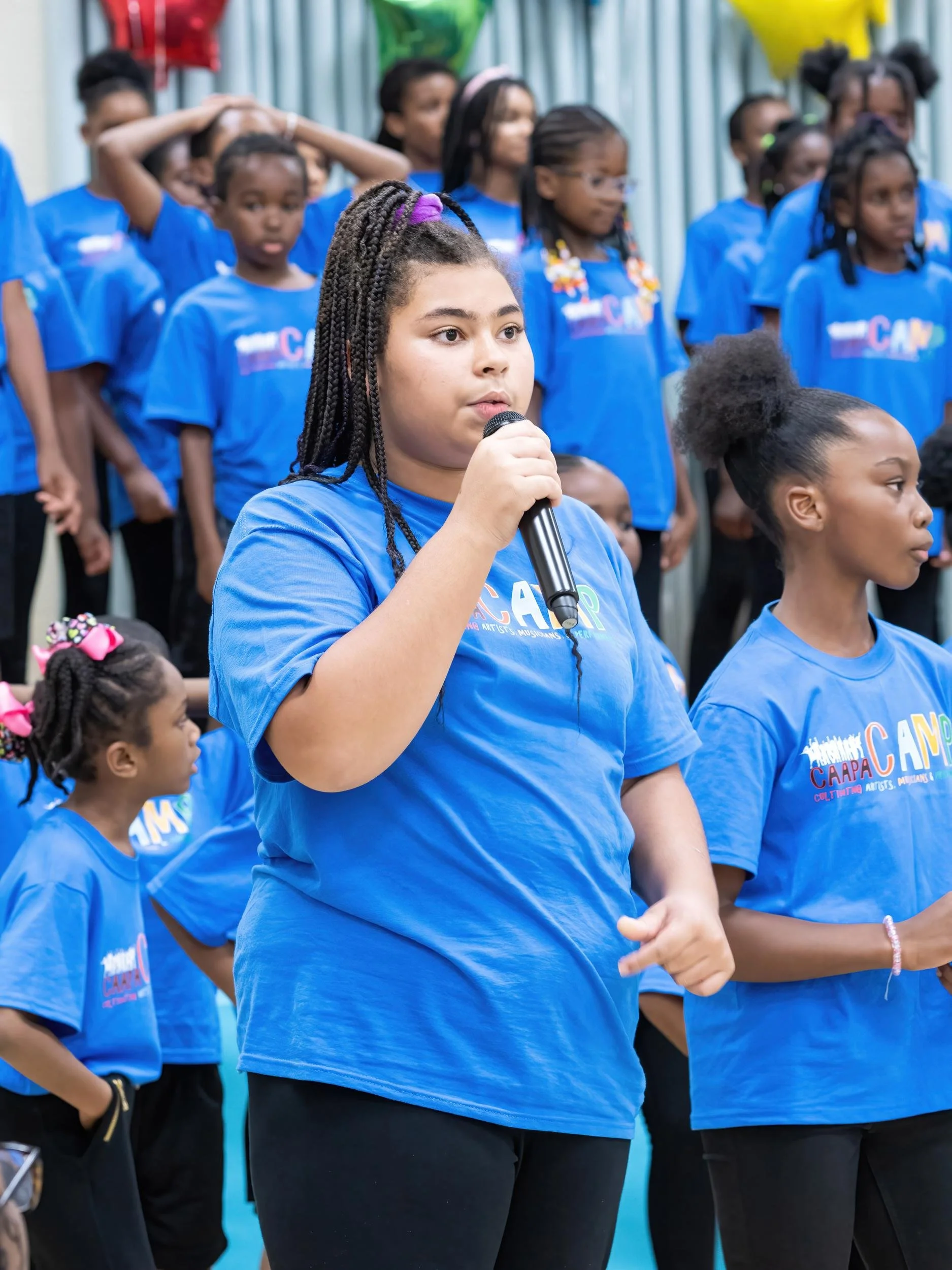 A young girl with braided hair holding a microphone, standing among a group of children wearing blue shirts at an outdoor event.
