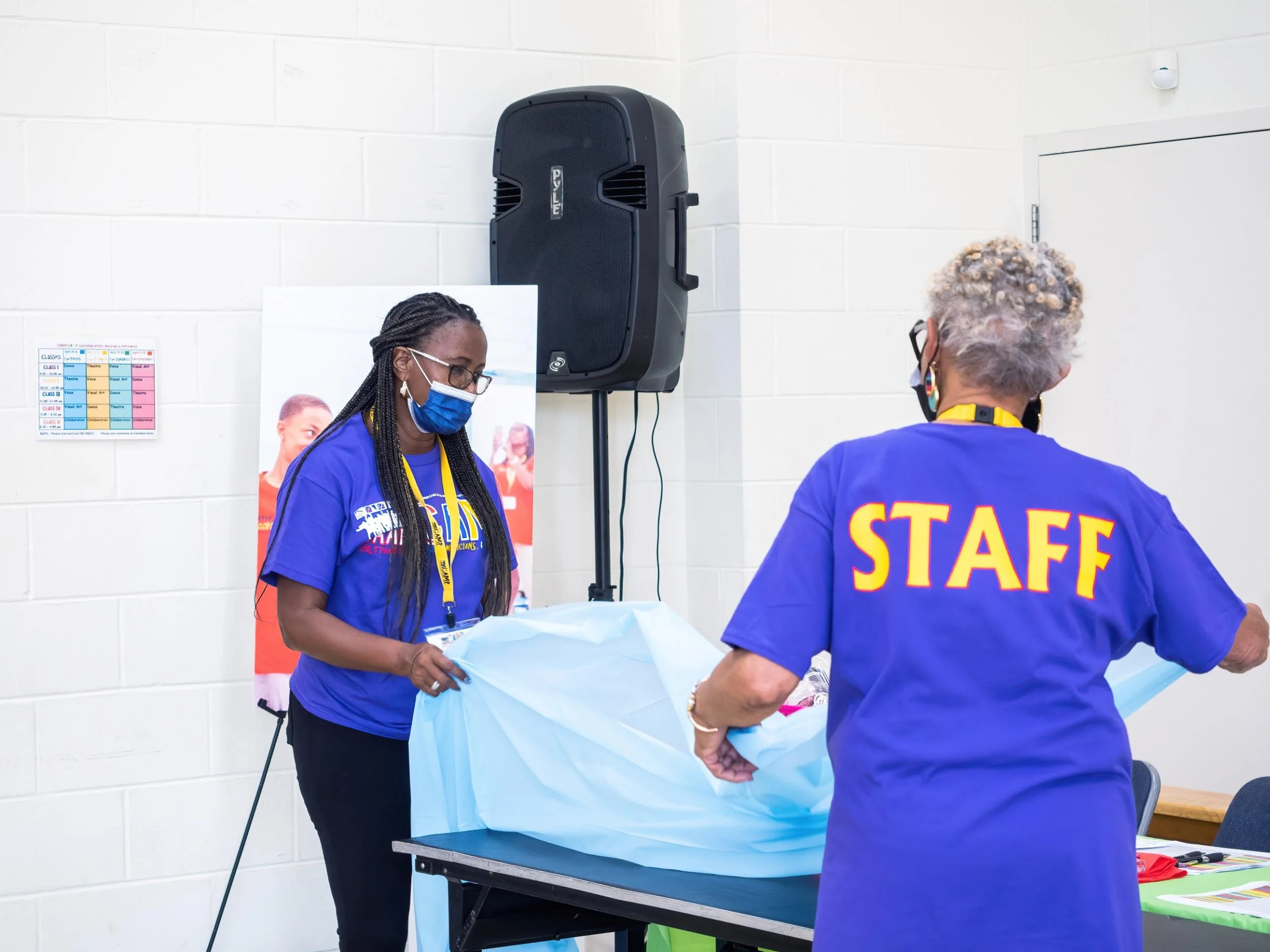 Two women, one with braided hair and the other with curly gray hair, wearing purple shirts, one with 'STAFF' printed on the back, working at a table with a large blue cloth, in a room with white cinder block walls and a large speaker in the backgroun