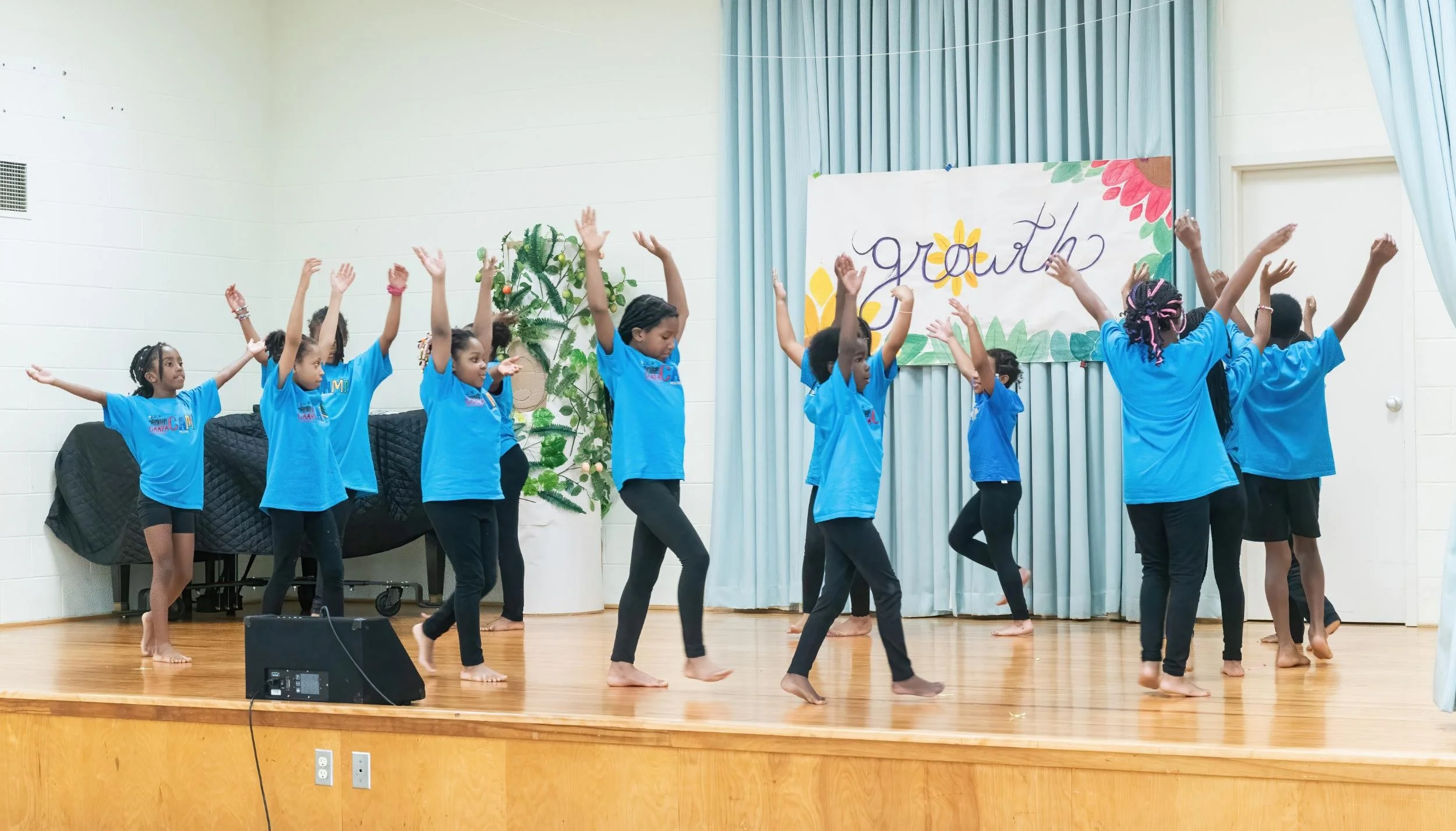 Children in matching blue shirts performing a dance on a stage with a sign that says 'growth' and decorative plants in the background.