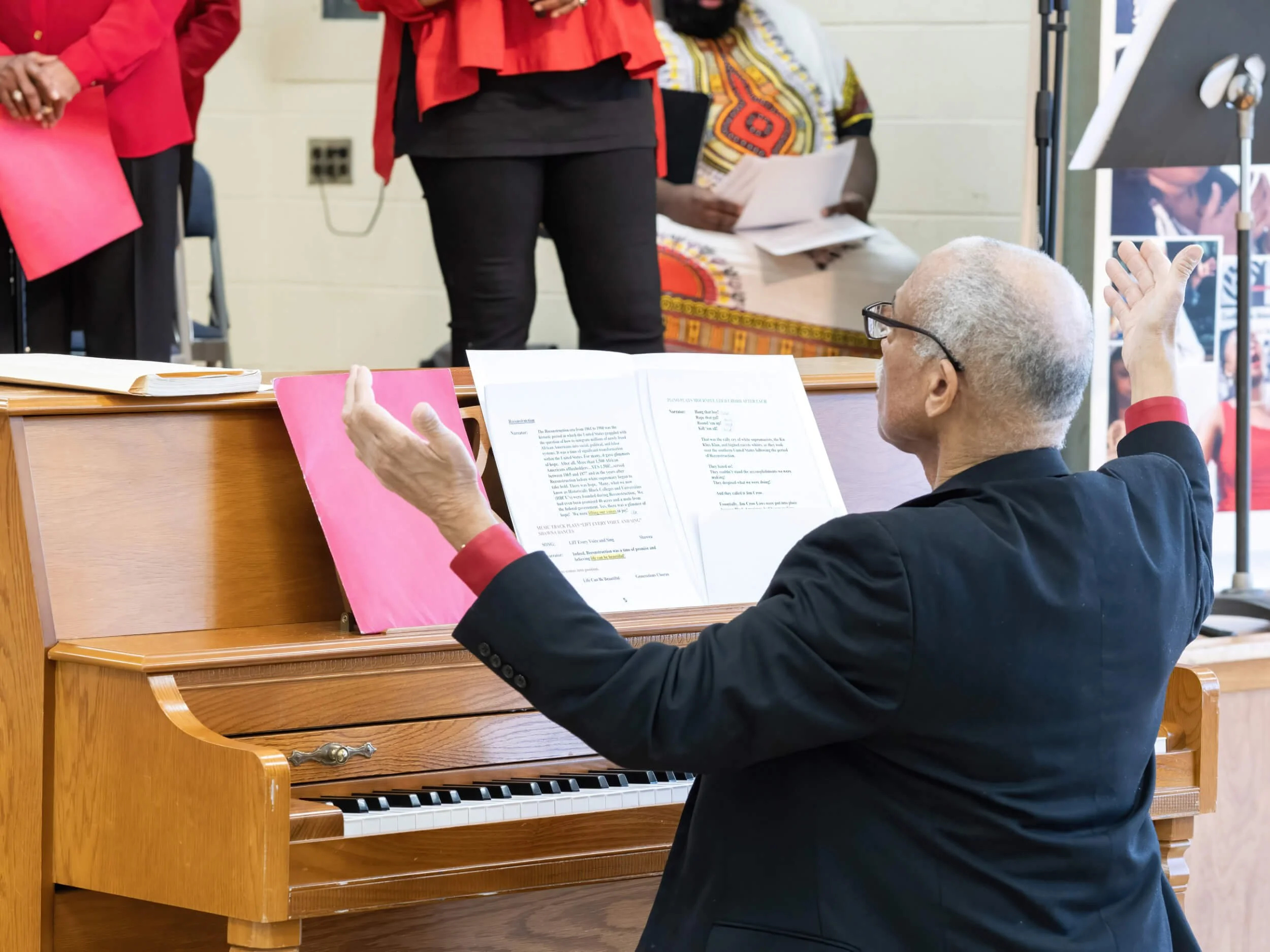 An elderly man with glasses and gray hair, dressed in a black suit, is sitting at a piano with sheet music in front of him. He has his arms raised and appears to be conducting or leading a singing group during a performance or rehearsal. Several peop