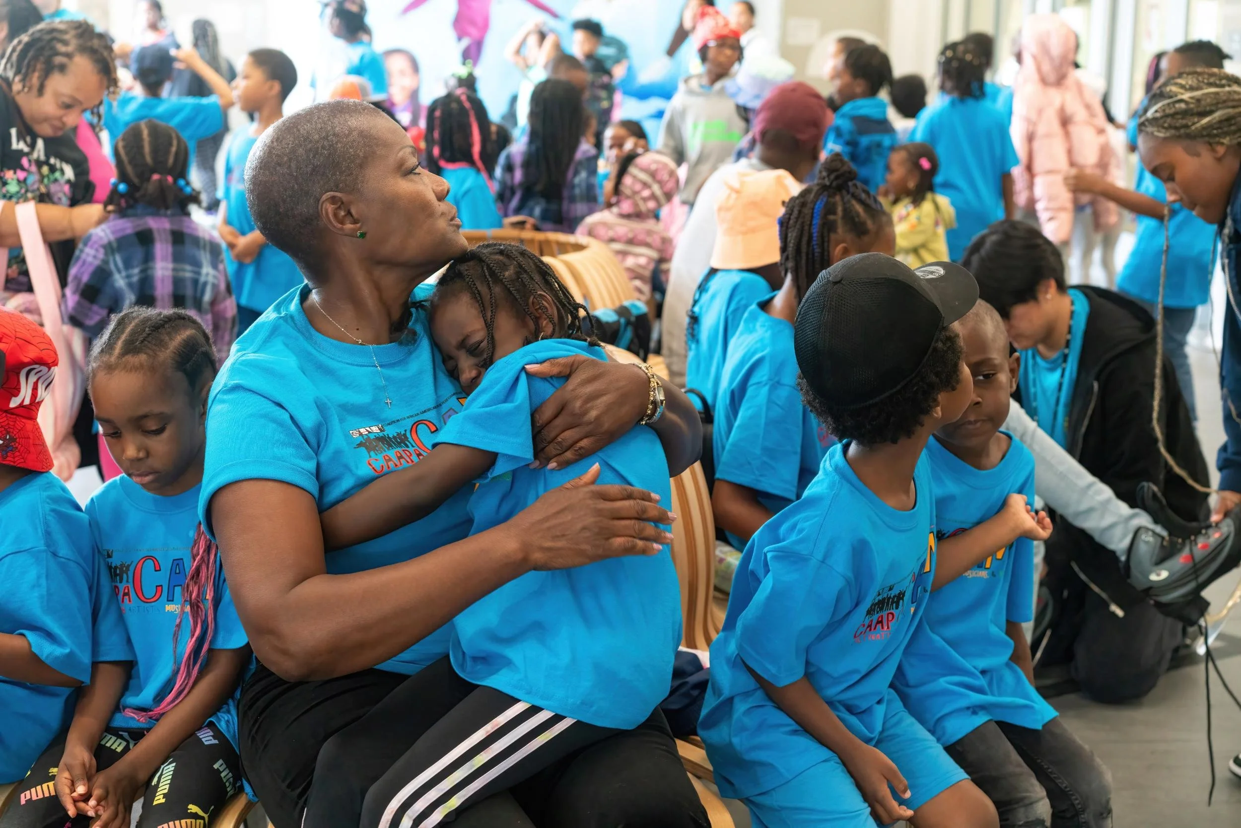 An African American woman with short hair, wearing a blue t-shirt, is sitting down and hugging a child who is resting their head on her shoulder. The woman appears emotional, as do some children around her in a busy indoor setting, possibly a group e