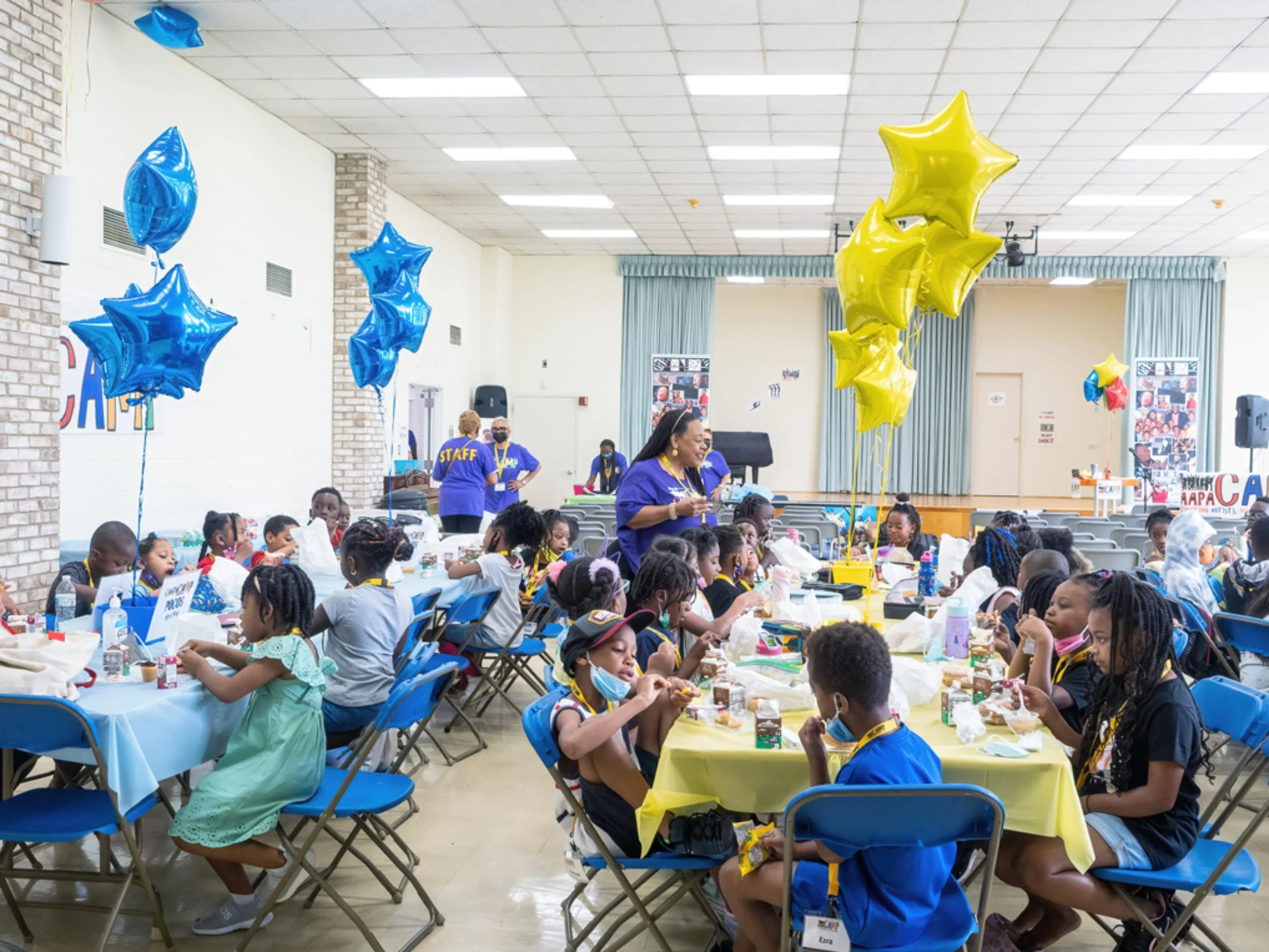 Children and staff at a birthday party or celebration in a large hall, decorated with blue and yellow star-shaped balloons, some children sitting at tables with food and drinks.