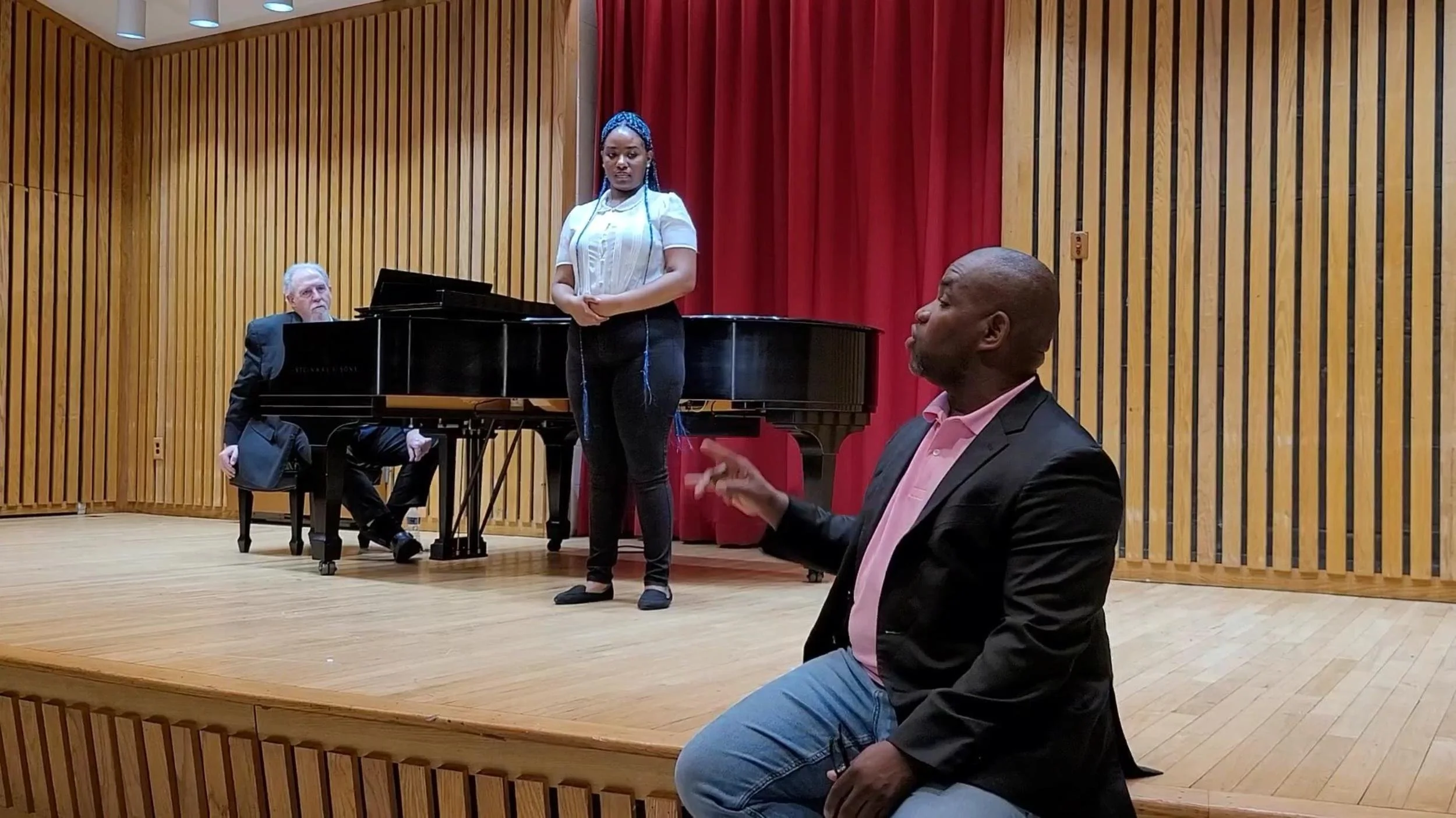 A woman with braided hair stands on a stage in front of a red curtain, with a man playing the piano behind her and another man sitting on the edge of the stage, speaking or gesturing.