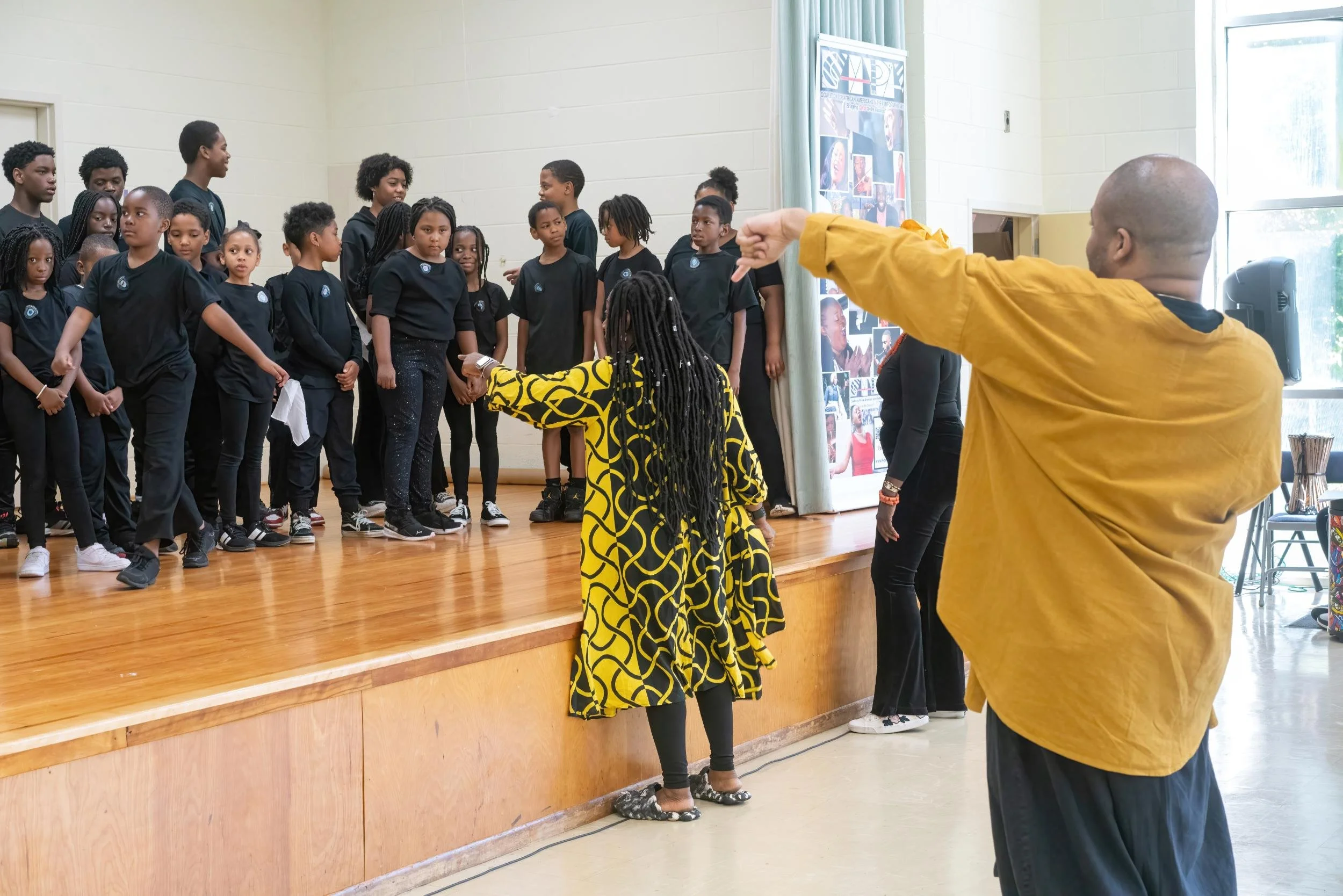 Children rehearsing on stage with two adults overseeing, in a bright room with large windows and a display board.