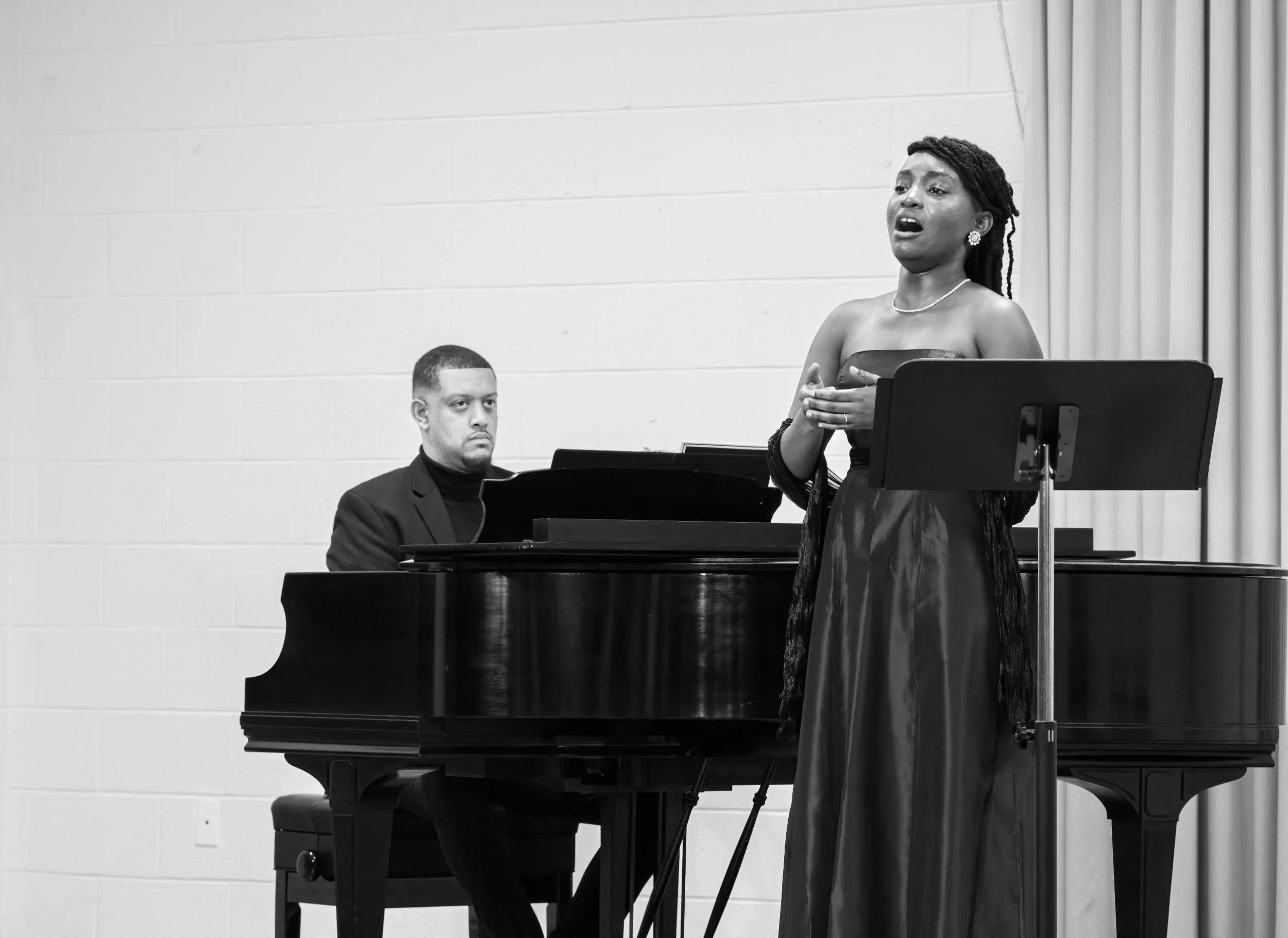 A woman singing with a sheet music stand in front of her, and a man playing a grand piano beside her, both performing on stage in black and white.