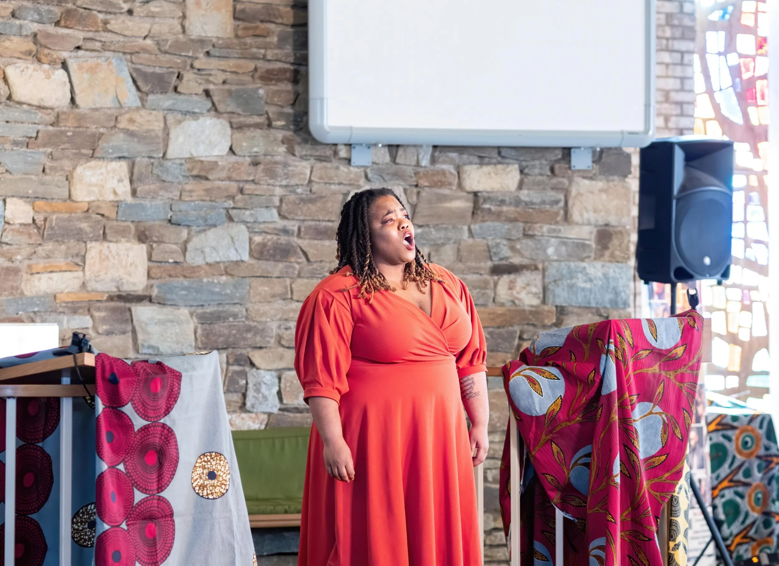 A woman with curly hair and wearing a red dress is singing or speaking passionately in front of a stone wall, with colorful fabric-covered objects around her and a microphone on a stand nearby.