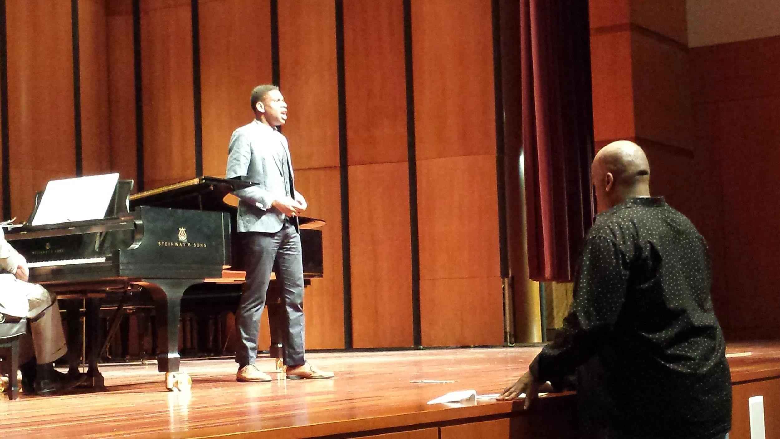 A person stands on stage, speaking or singing, next to a grand piano, with an audience member watching from the front row.