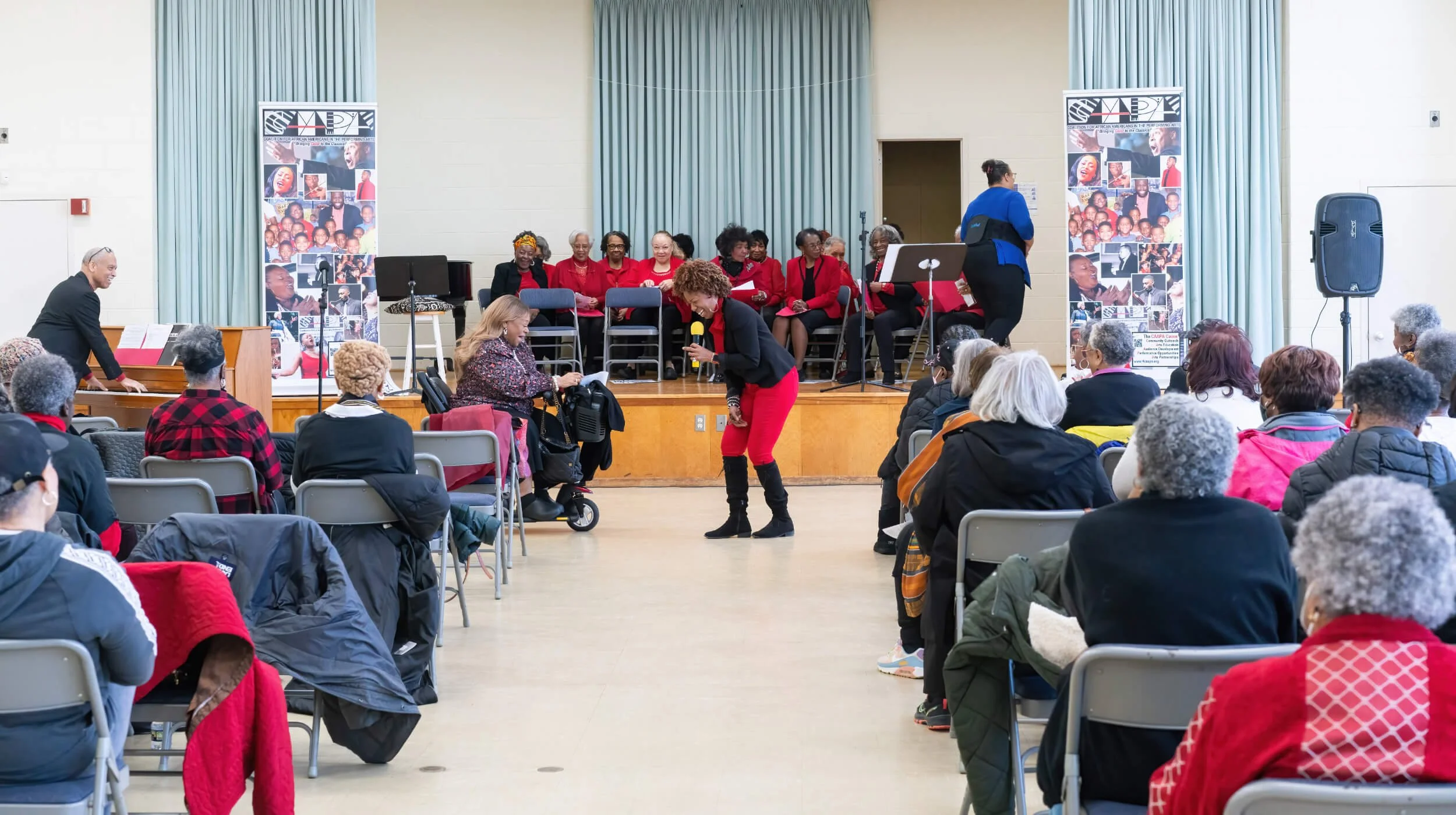 An indoor community event with a diverse group of elderly people seated in rows, a woman with curly hair singing into a microphone, a pianist playing, and a choir dressed in red sweaters on the stage with banners and posters.