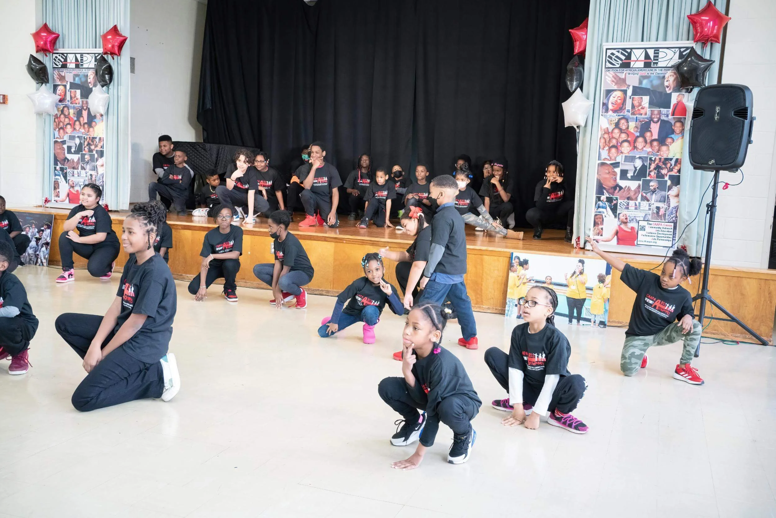 Children performing on stage and in the floor area, some kneeling and some sitting, during a dance recital or performance at a school or community center.