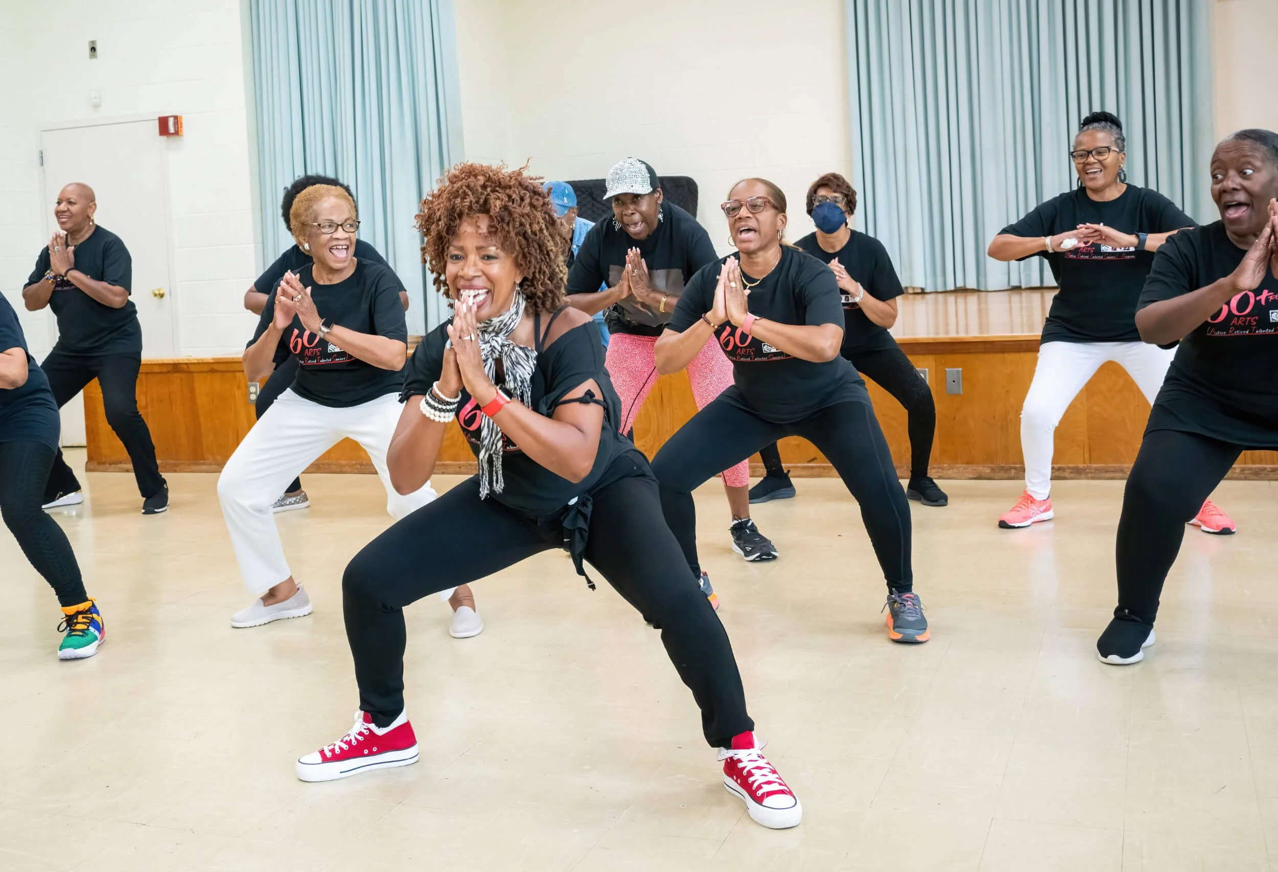 A group of women participating in a dance class, smiling and wearing black shirts, in a room with wooden floors and teal curtains.