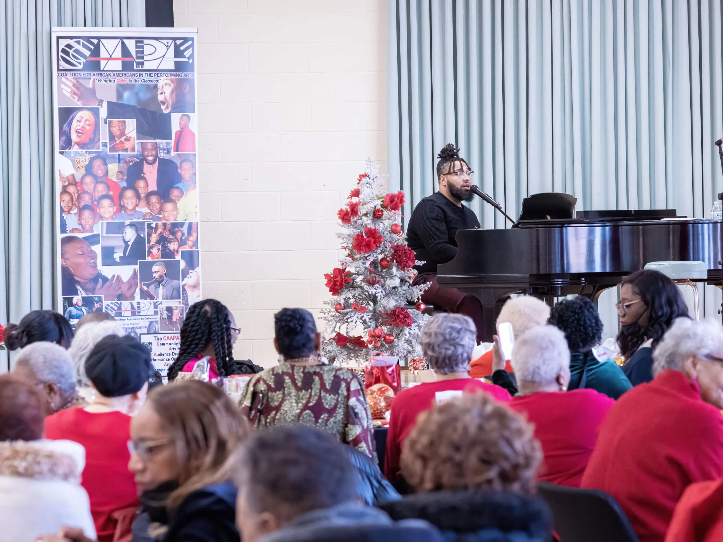 A man with glasses and dreadlocks singing at a black grand piano during a holiday event for a diverse audience of elderly and middle-aged people, with a Christmas tree beside him and a banner on the left side of the stage.