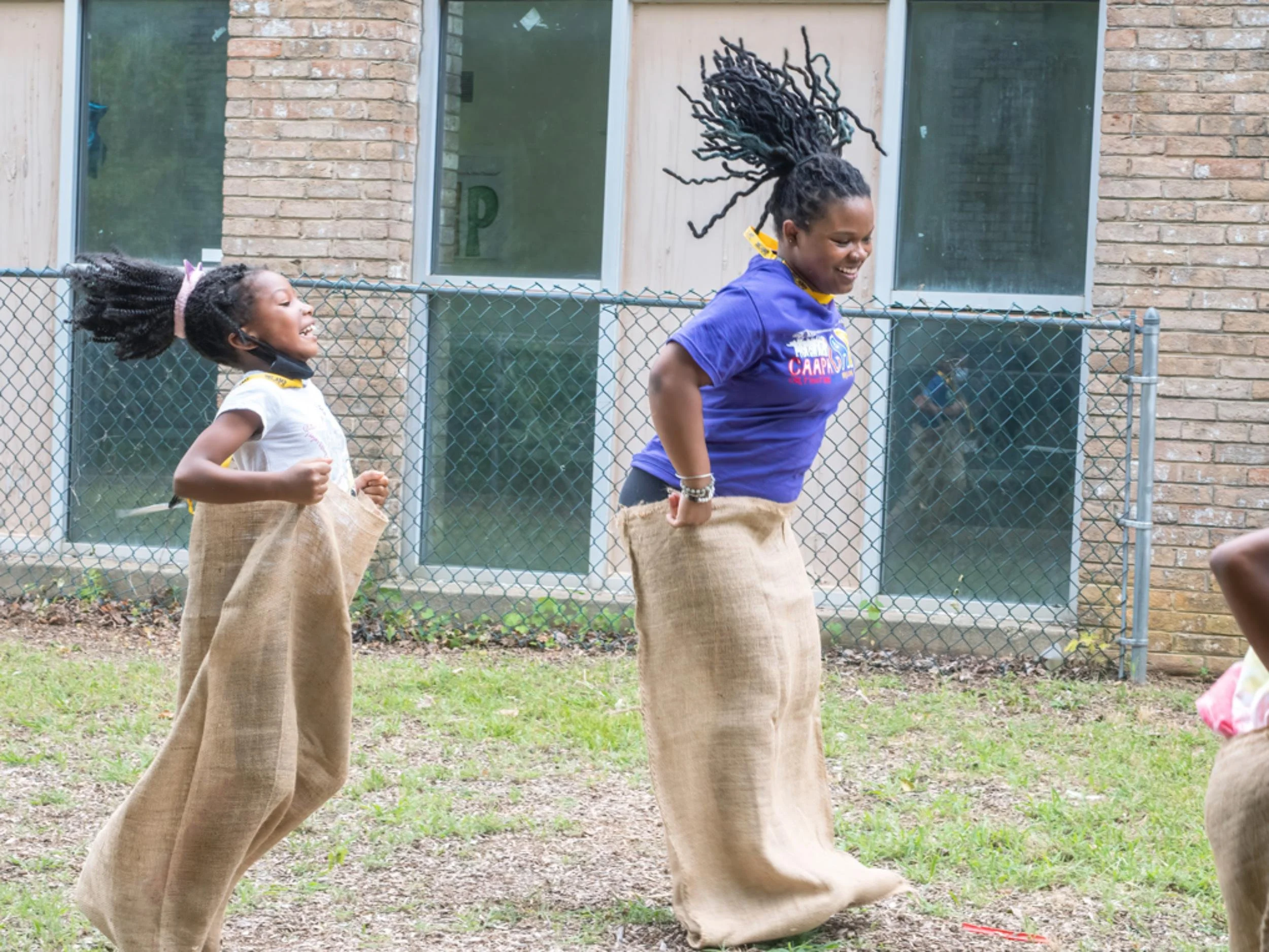Two young girls with dreadlocks participating in sack races outdoors, smiling and laughing, near a chain-link fence with a brick building and windows in the background.