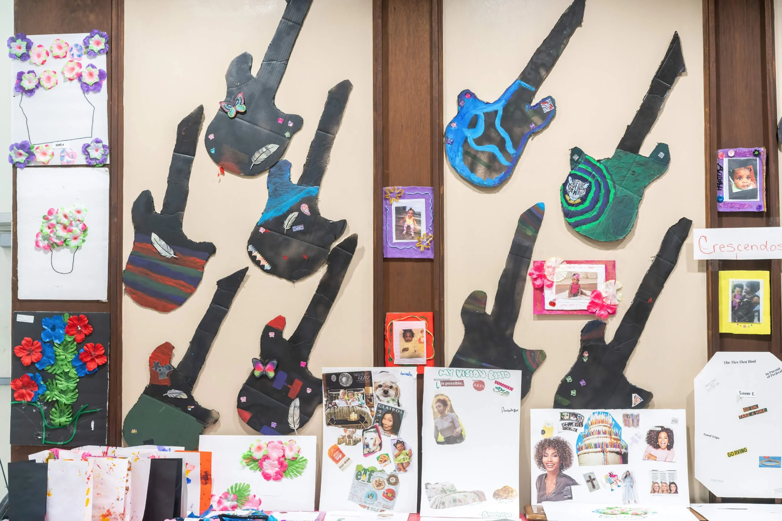 Display of various children’s artwork and photographs arranged on a wall and table, including paper flowers, handmade crafts, and framed photos of kids.