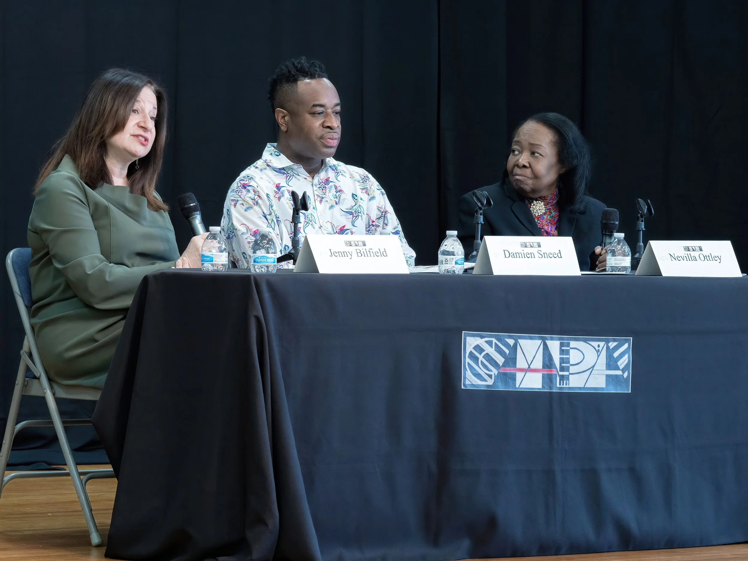Panel discussion with four people at a table with black tablecloth and microphones. The panel has name cards reading Jenny Bilfield, Damien Sneed, and Nevilla Ottley.