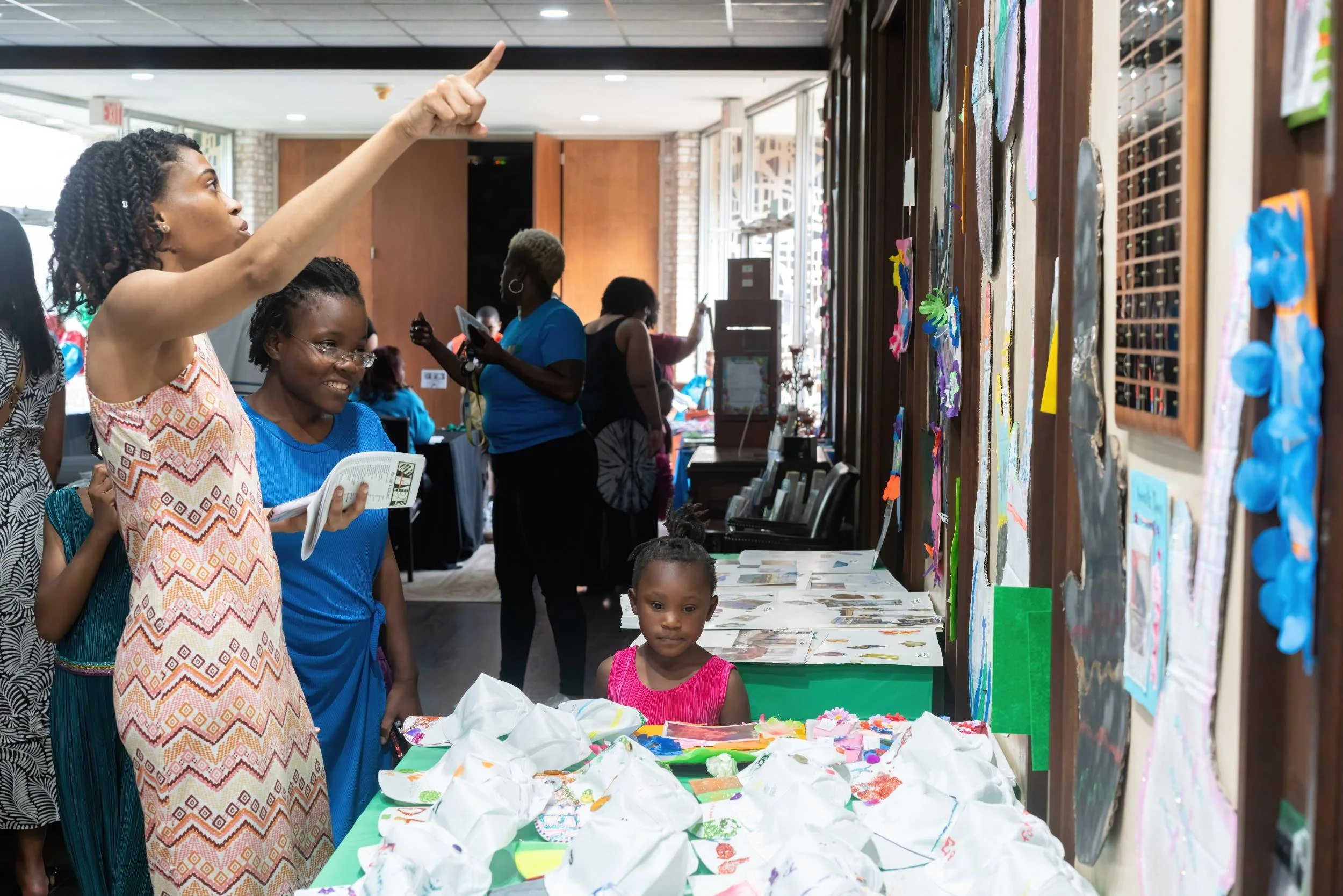 A diverse group of women and children are at an indoor art event, viewing and discussing colorful artwork and crafts displayed on a wall and tables.