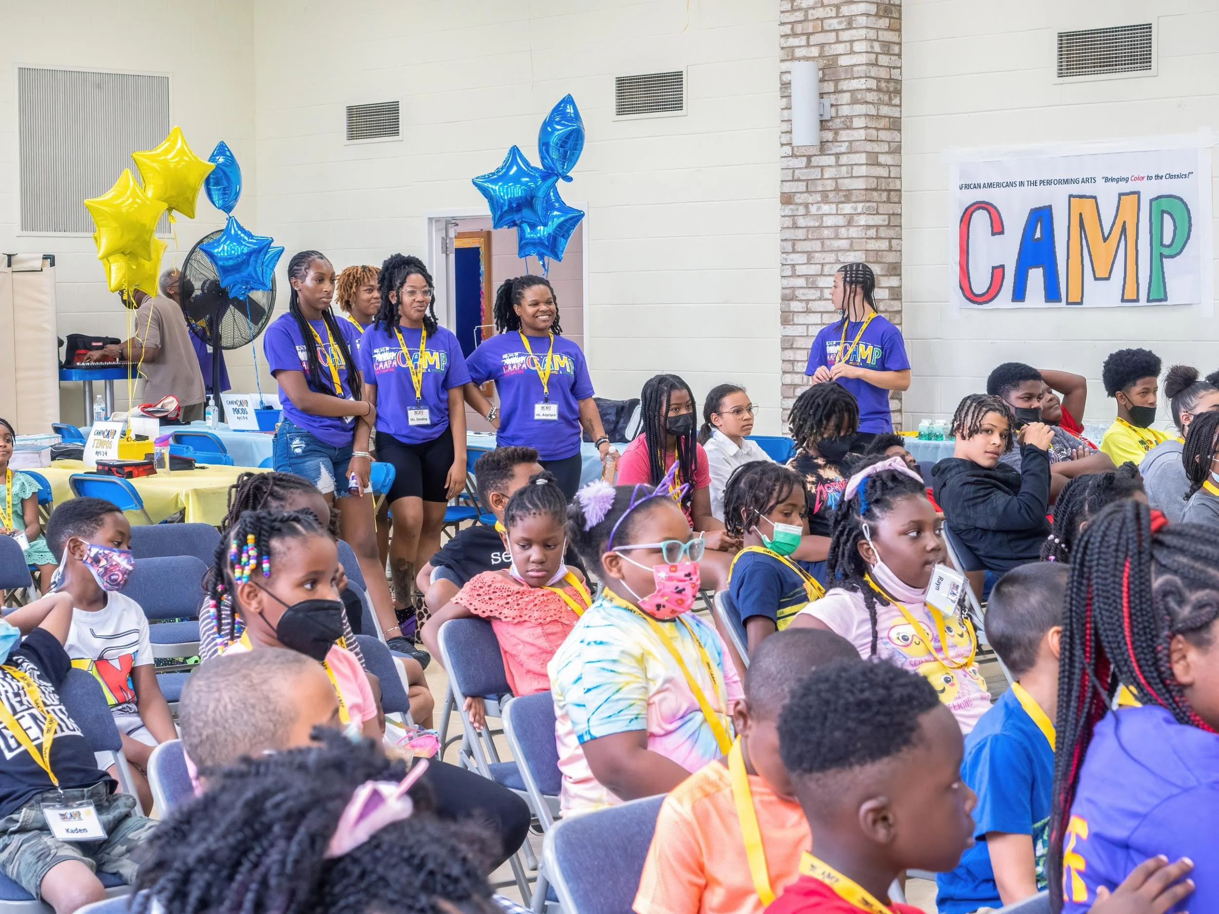 Children and young adults attending a camp event indoors, sitting on chairs, many wearing masks, some standing near a table with balloons, a large colorful sign reading 'CAMP' on the wall.