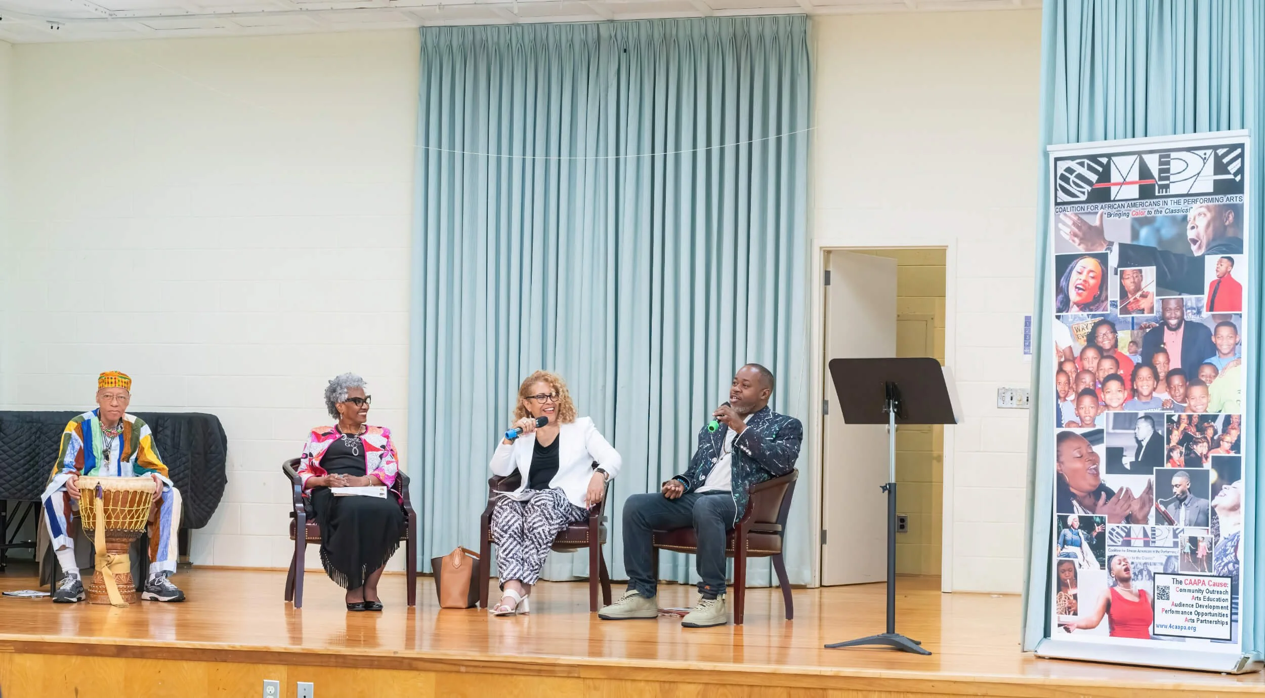 Four people sitting on stage engaging in a discussion, with one woman holding a microphone, and a large poster on the right side featuring images of various performers and text about an organization supporting performing arts and education.