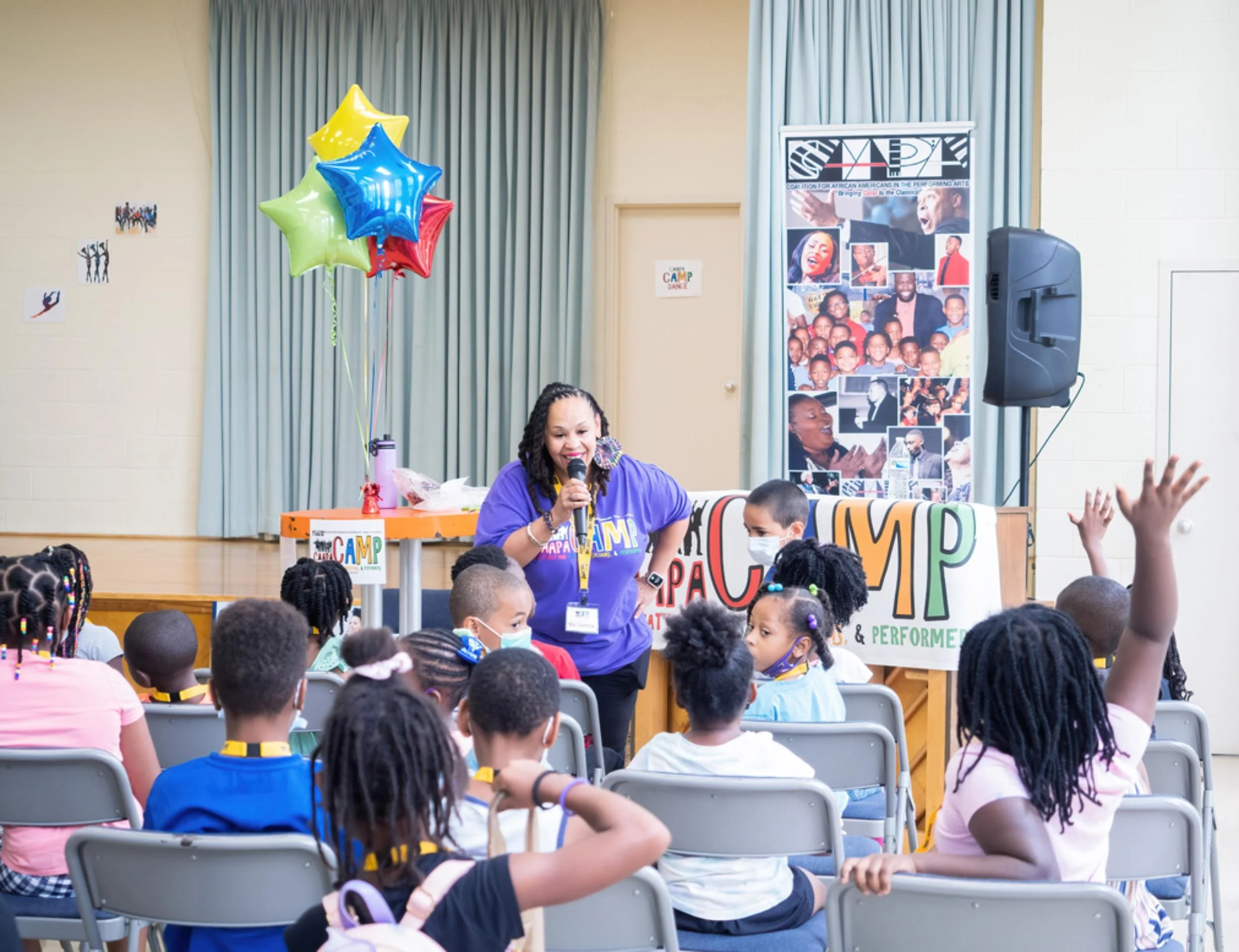 A woman holding a microphone speaking to a group of children at a camp event, with children raising their hands, balloons, and a banner in the background.