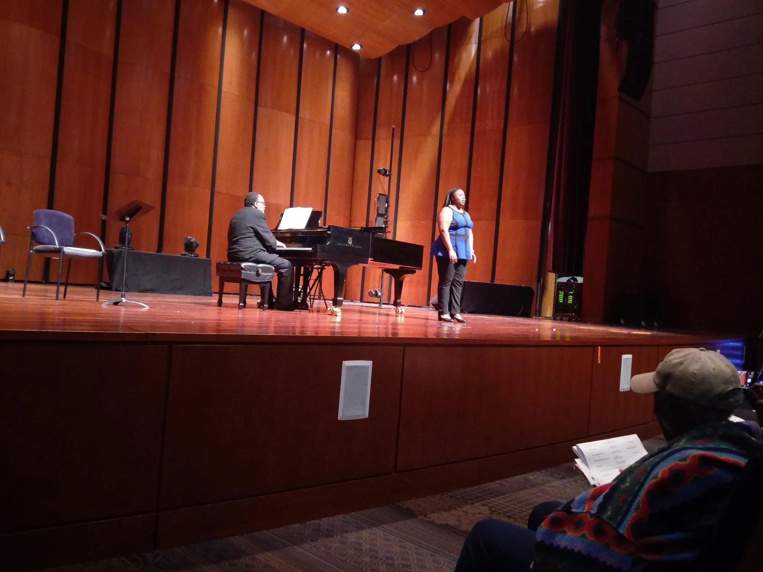 Music rehearsal on stage: a woman singing standing, a man playing a grand piano, audience member reading sheet music.