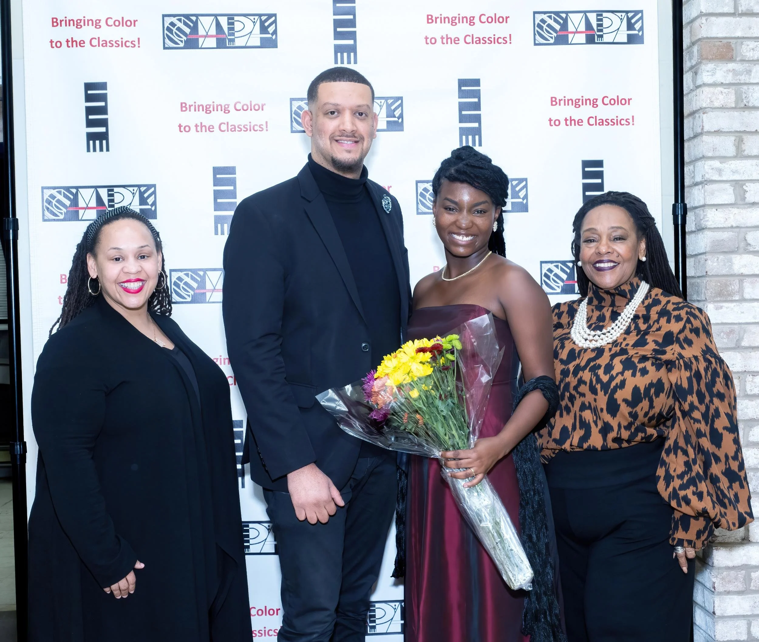 Group of four women and one man at a formal event, with a woman in the center holding a bouquet of flowers, standing in front of a backdrop with logos and text that reads 'Bringing Color to the Classics!'.