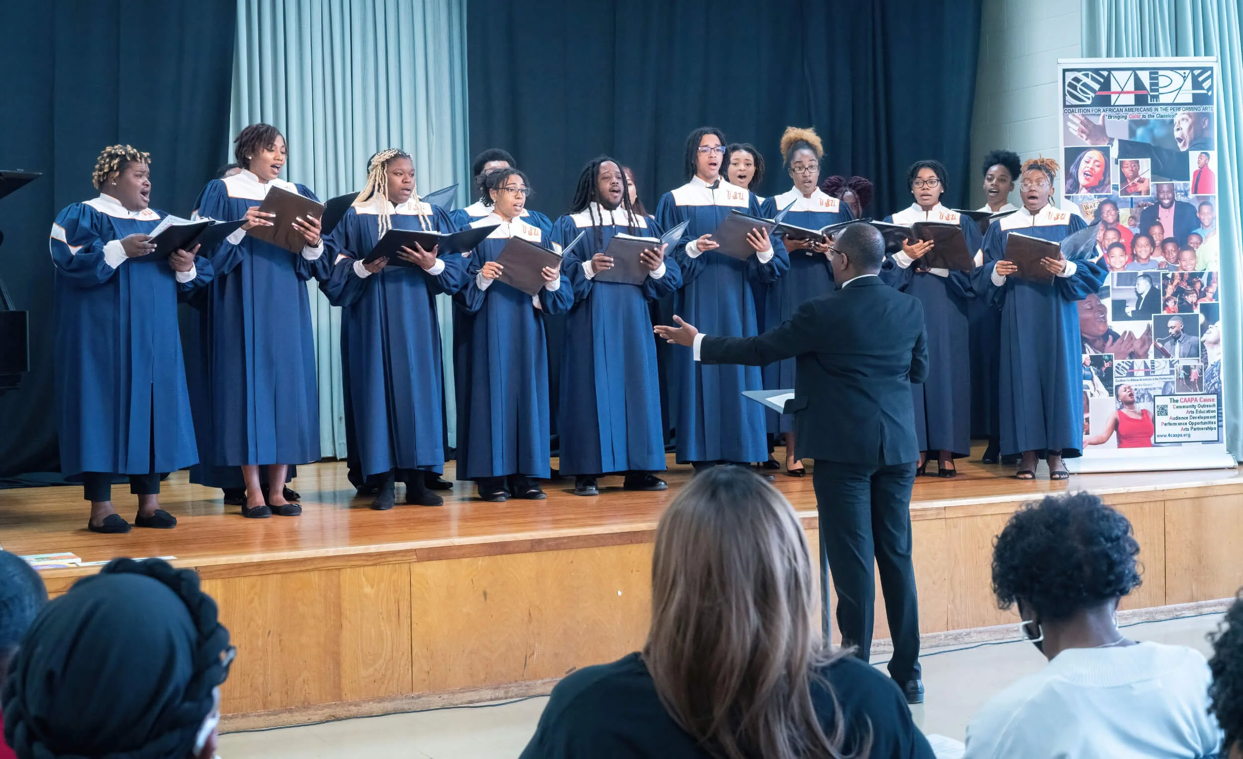 Choir of African American women singing on stage, directed by a conductor in a black suit, during a performance at an indoor event, with an audience watching.