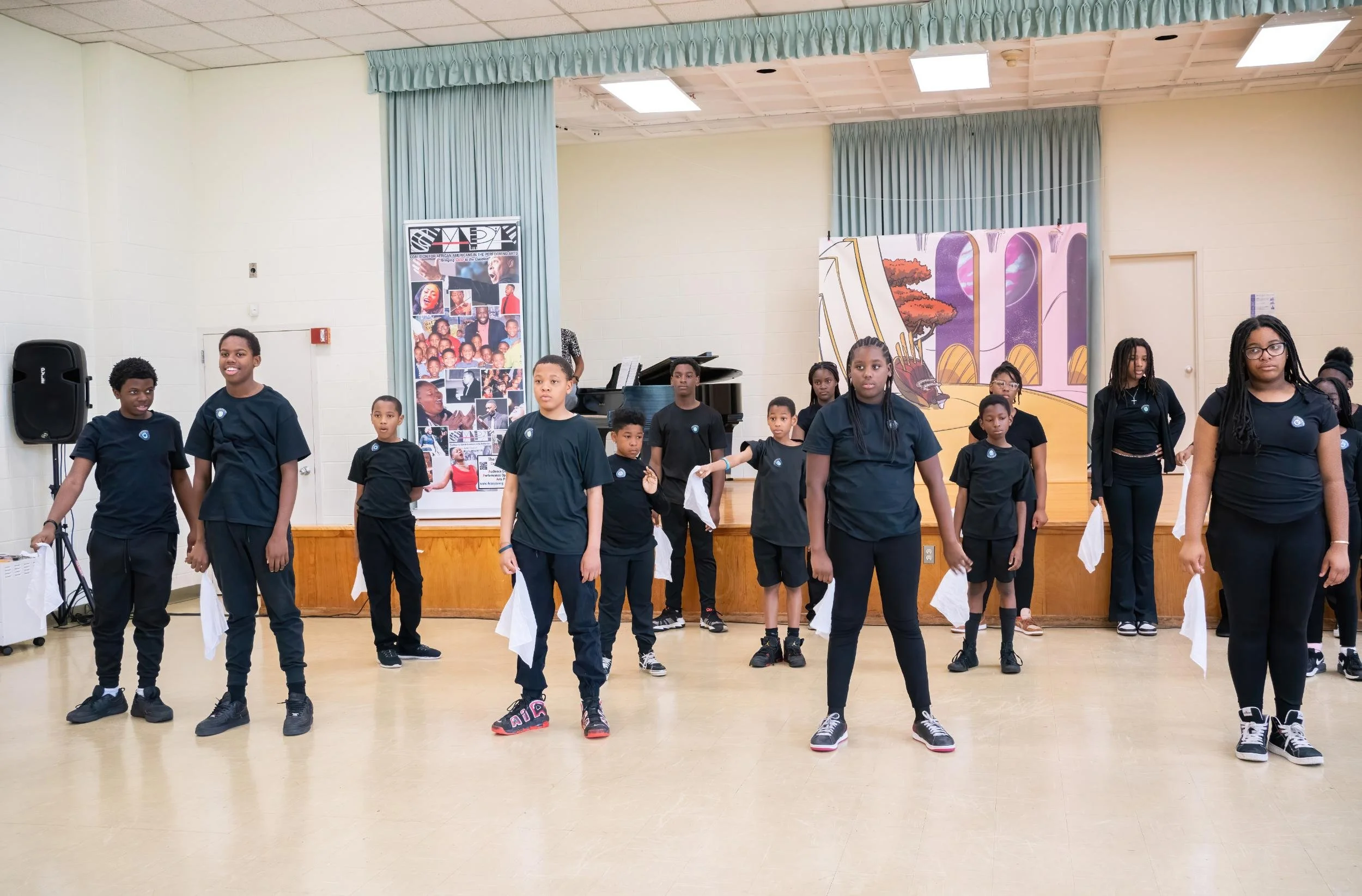 Children performing a dance in a school gymnasium or auditorium, dressed in black T-shirts and pants, with some holding white cloths, standing in front of a decorated stage with a backdrop and displayed banner.
