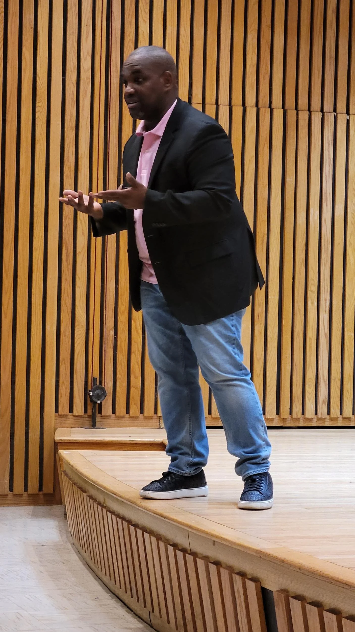 A man in a black blazer, pink shirt, blue jeans, and sneakers standing on a wooden stage with a wood-paneled background, giving a speech or presentation.