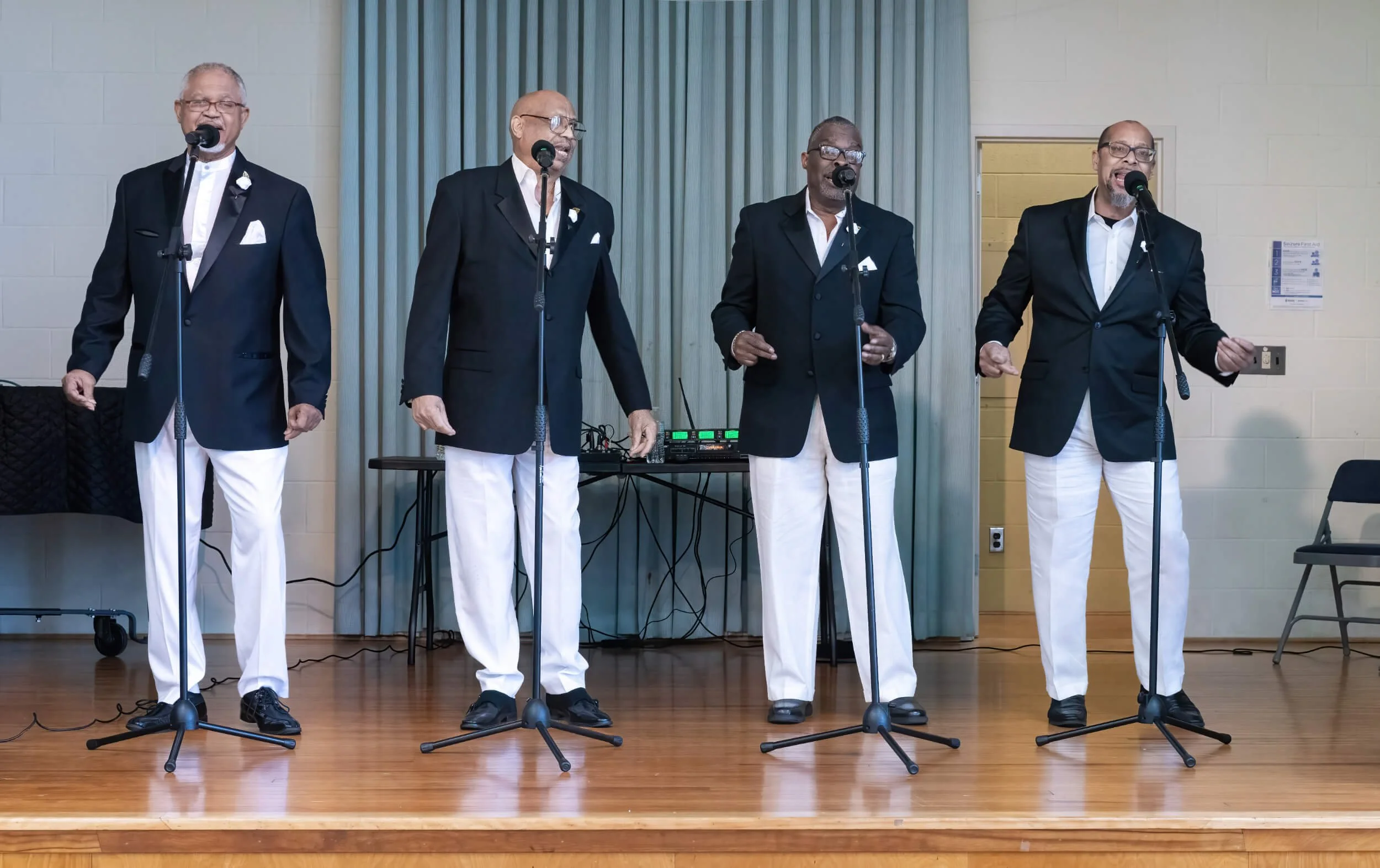 Four men singing into microphones on a stage, wearing black blazers, white pants, and white shirts, with a DJ setup behind them.
