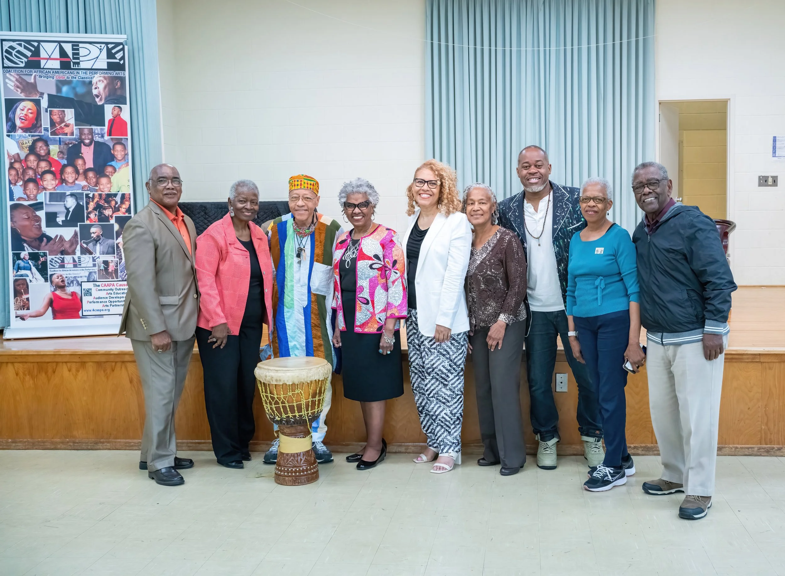 Group of ten diverse people standing in a line indoors, smiling at the camera, with a drum and a poster featuring photos of African American performers and events in the background.