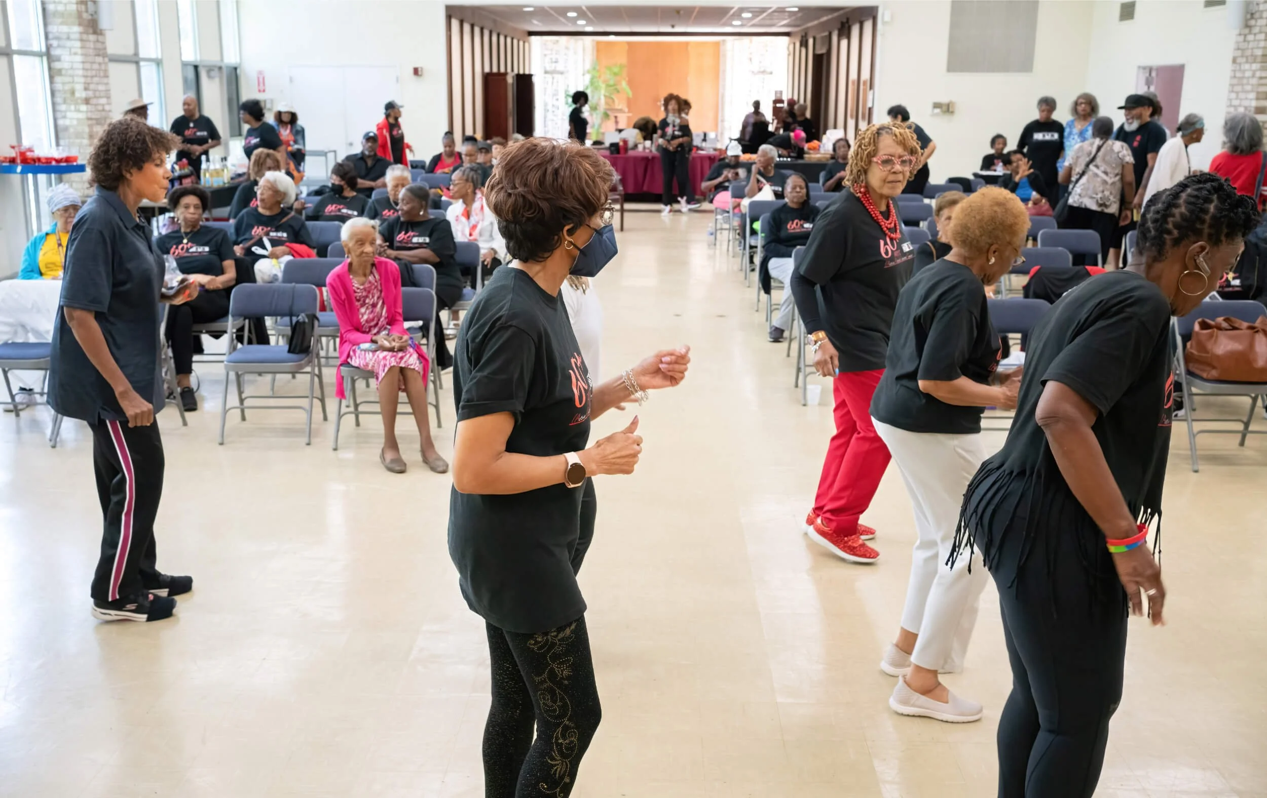 Women of various ages dancing and socializing in a large indoor space with seated attendees in the background.