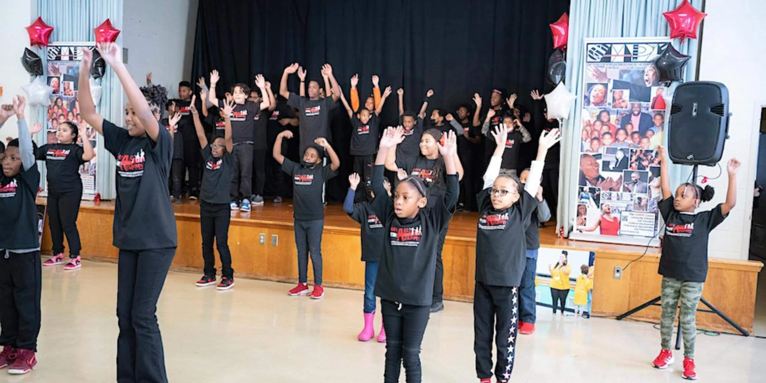 Children participating in a dance or exercise class in a school gymnasium with a stage, decorated with red, white, and black balloons, and posters of African American figures.
