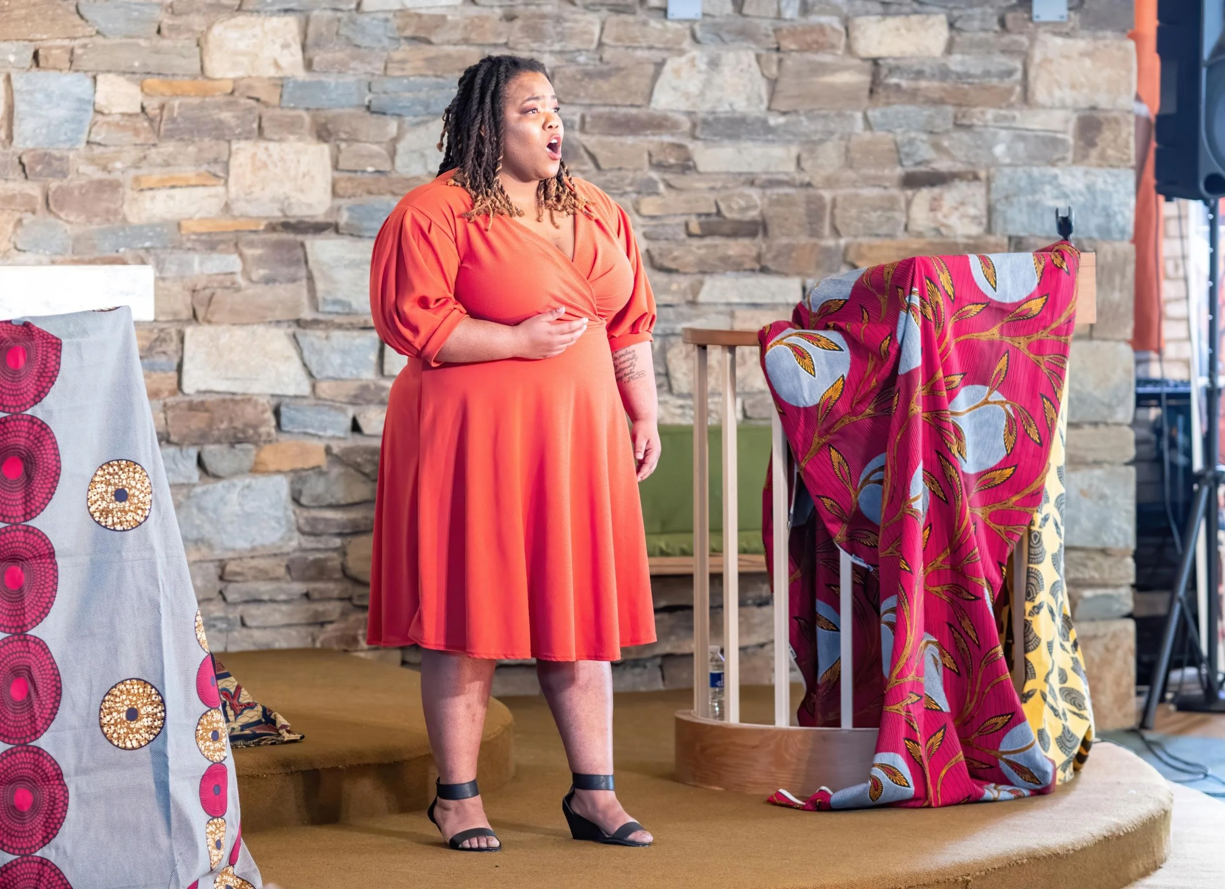 A woman singing on stage, wearing an orange dress, standing in front of a stone wall with colorful fabric draped over some structures.