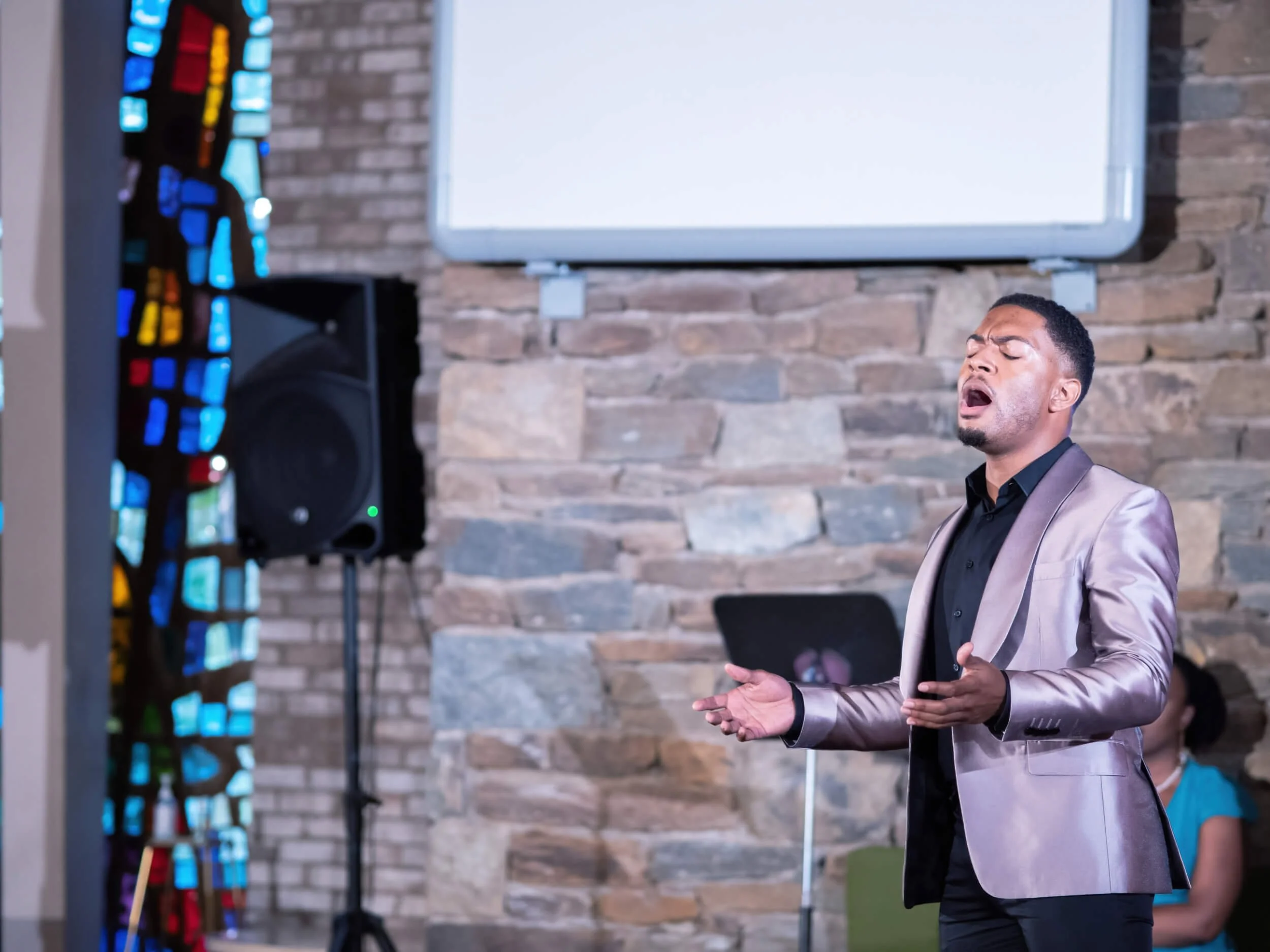Man singing passionately with eyes closed, wearing a shiny blazer and holding a microphone in a room with brick wall background, a fluorescent whiteboard, and audience members.