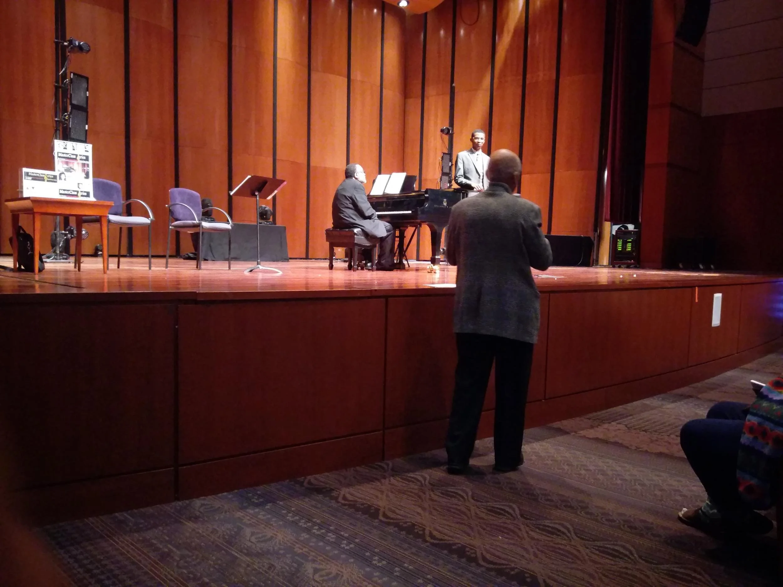 A jazz performance on stage with a pianist, a vocalist, and three empty chairs; an audience member standing near the stage in a dimly lit auditorium.