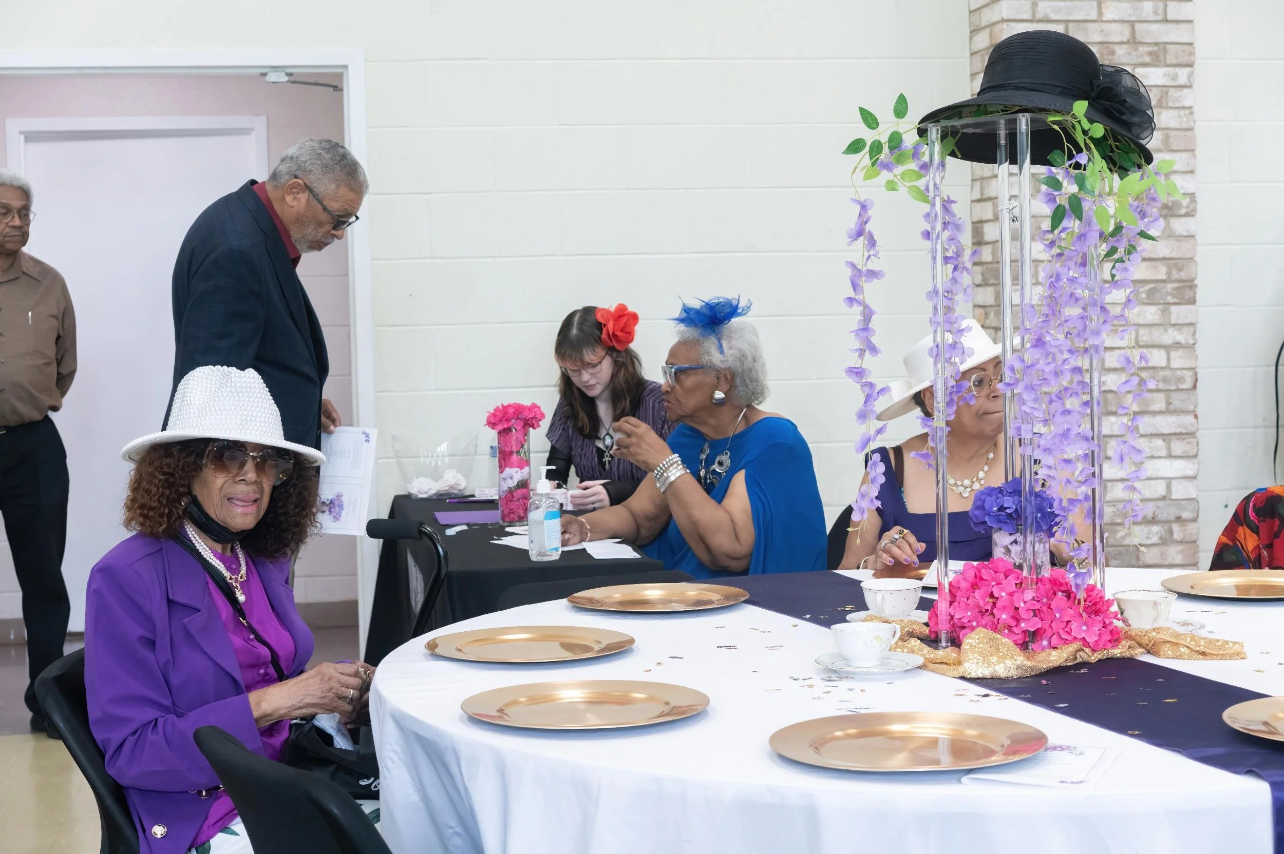 A social gathering with women dressed in colorful clothing, wearing hats, and sitting around a decorated table with purple and pink flowers, gold plates, and teacups.