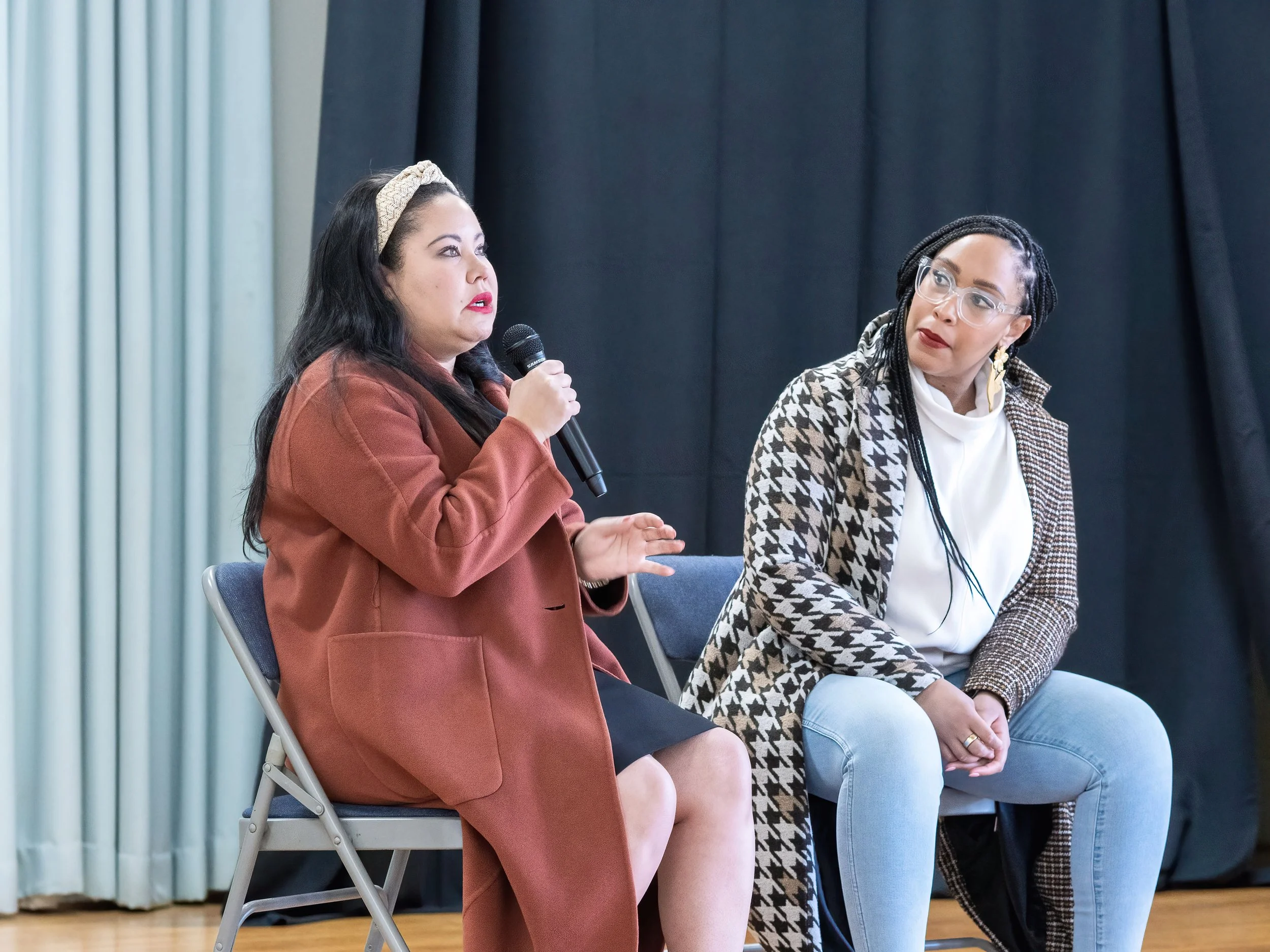 Two women sitting on stage, one holding a microphone and speaking, the other listening attentively, with a black curtain behind them.
