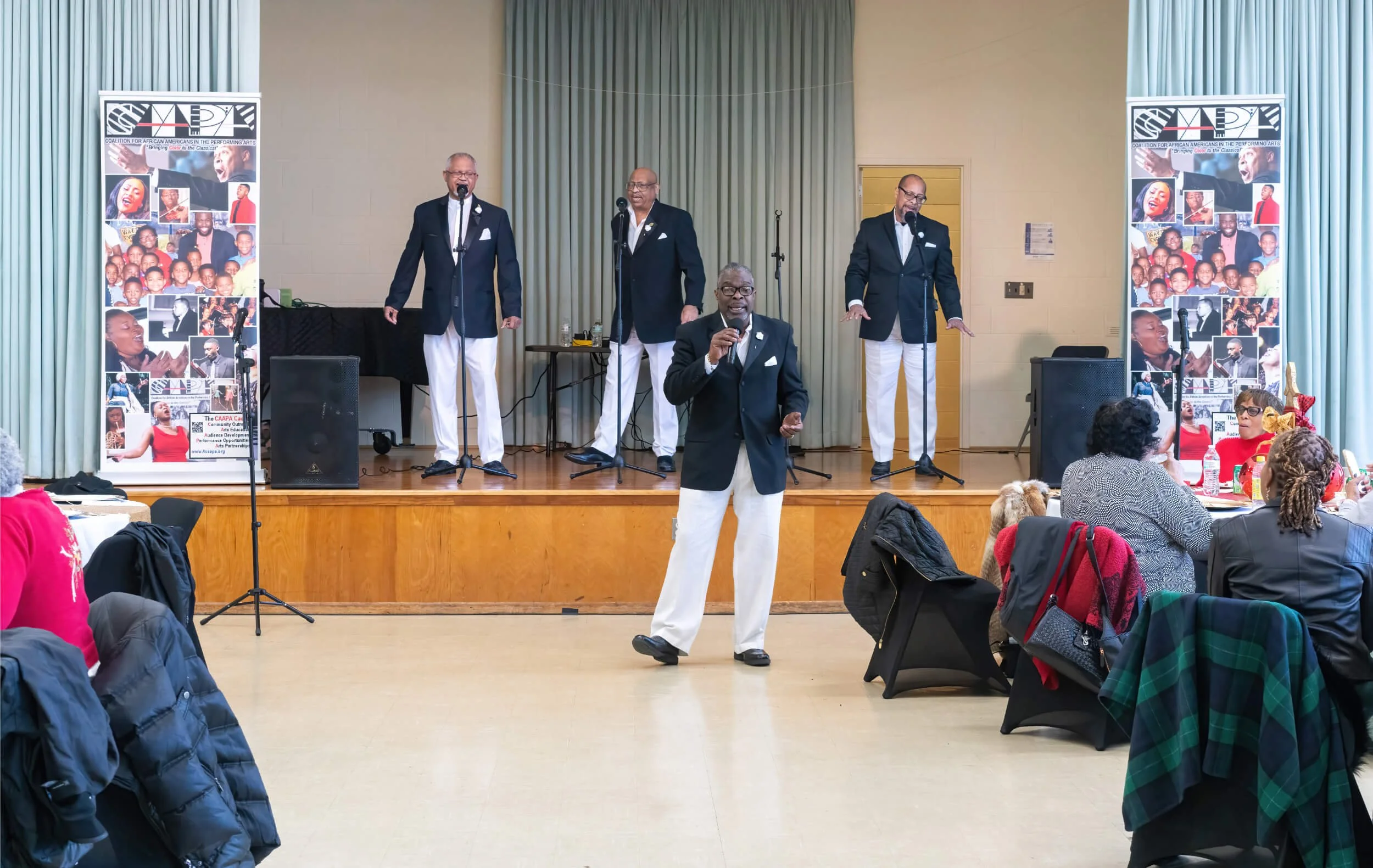 A group of five African American men dressed in black blazers, white pants, and ties, performing on a stage with microphones at a community event, with seated audience members watching.
