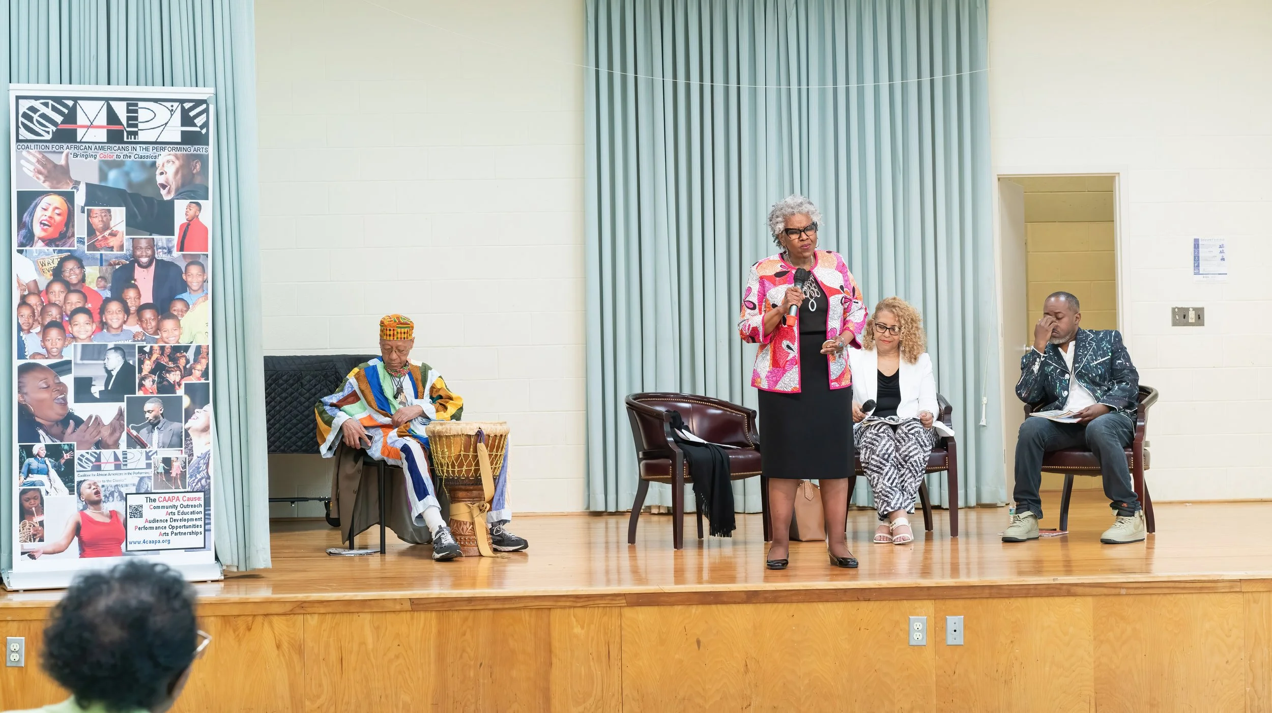 A panel discussion with four people on stage, including a woman speaking into a microphone, a woman with curly hair, a man in a patterned jacket, and an older man in traditional African attire with a drum, in front of a large banner promoting African