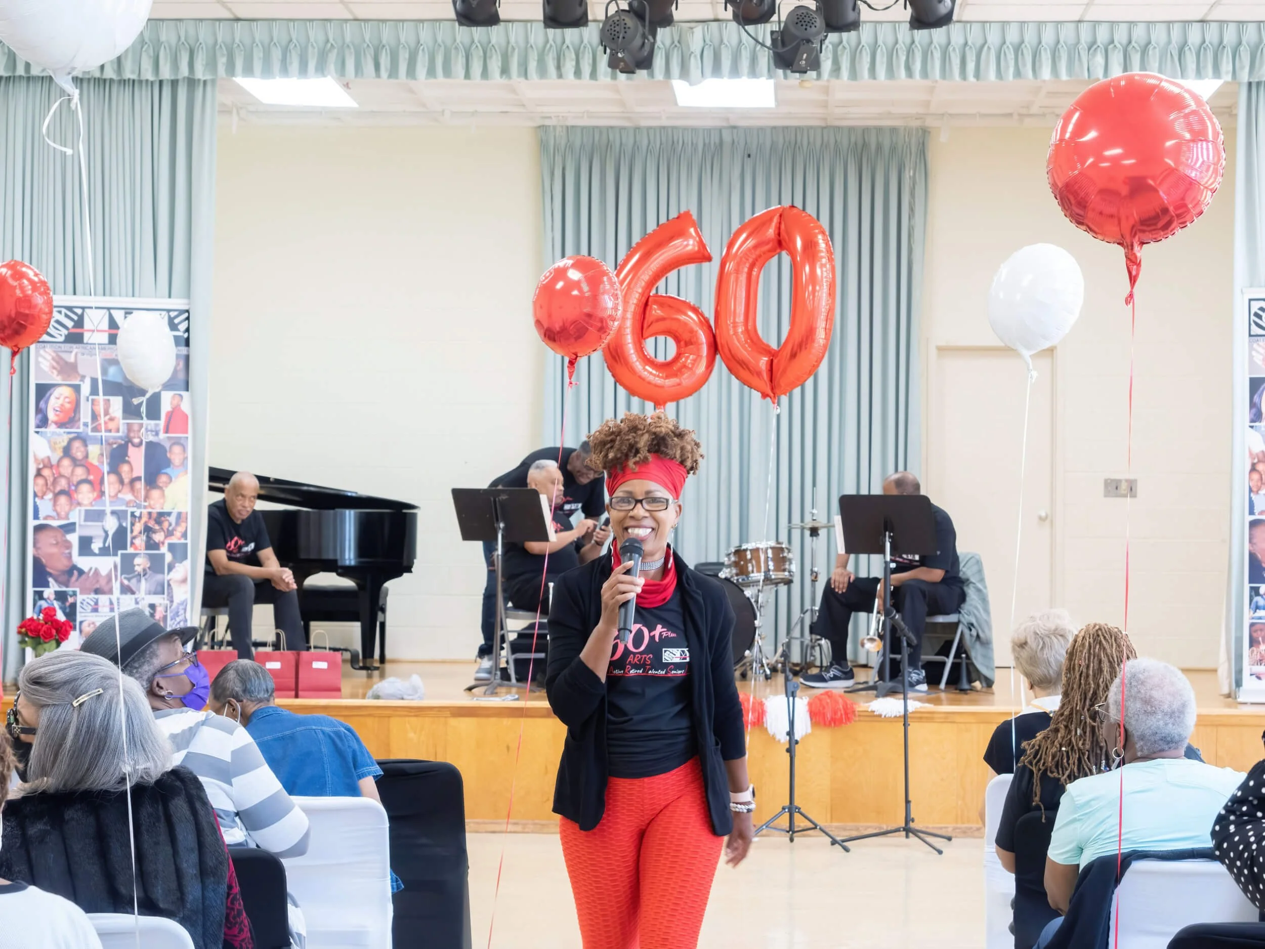 A woman smiles holding a microphone at a celebration with red and white balloons, including red balloons shaped as '60' and red and white balloons on strings. She stands in front of a stage with musical instruments, and an audience is seated facing h