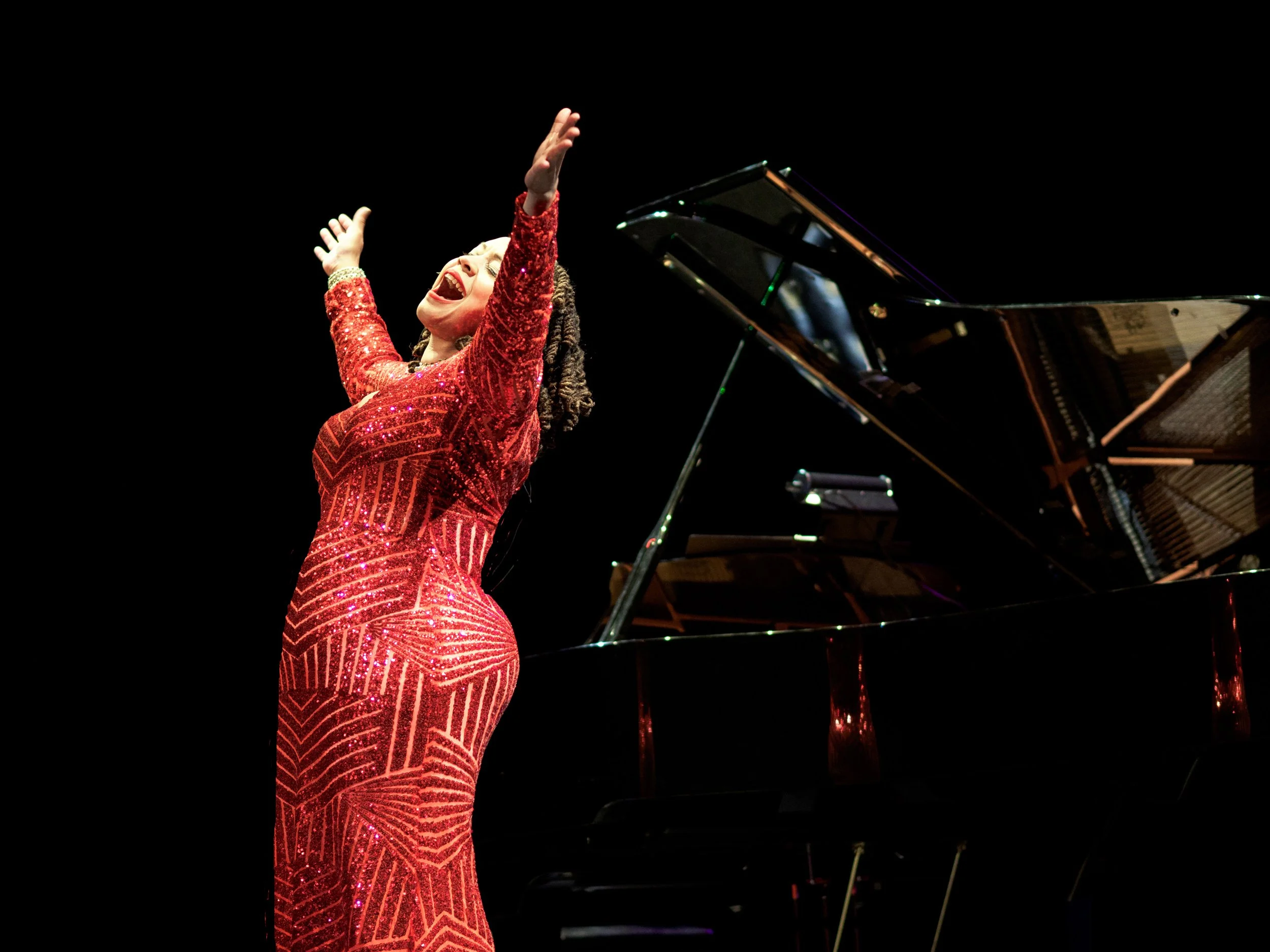 A woman in a red, sequined dress passionately singing on stage with her arms raised, beside a grand piano.