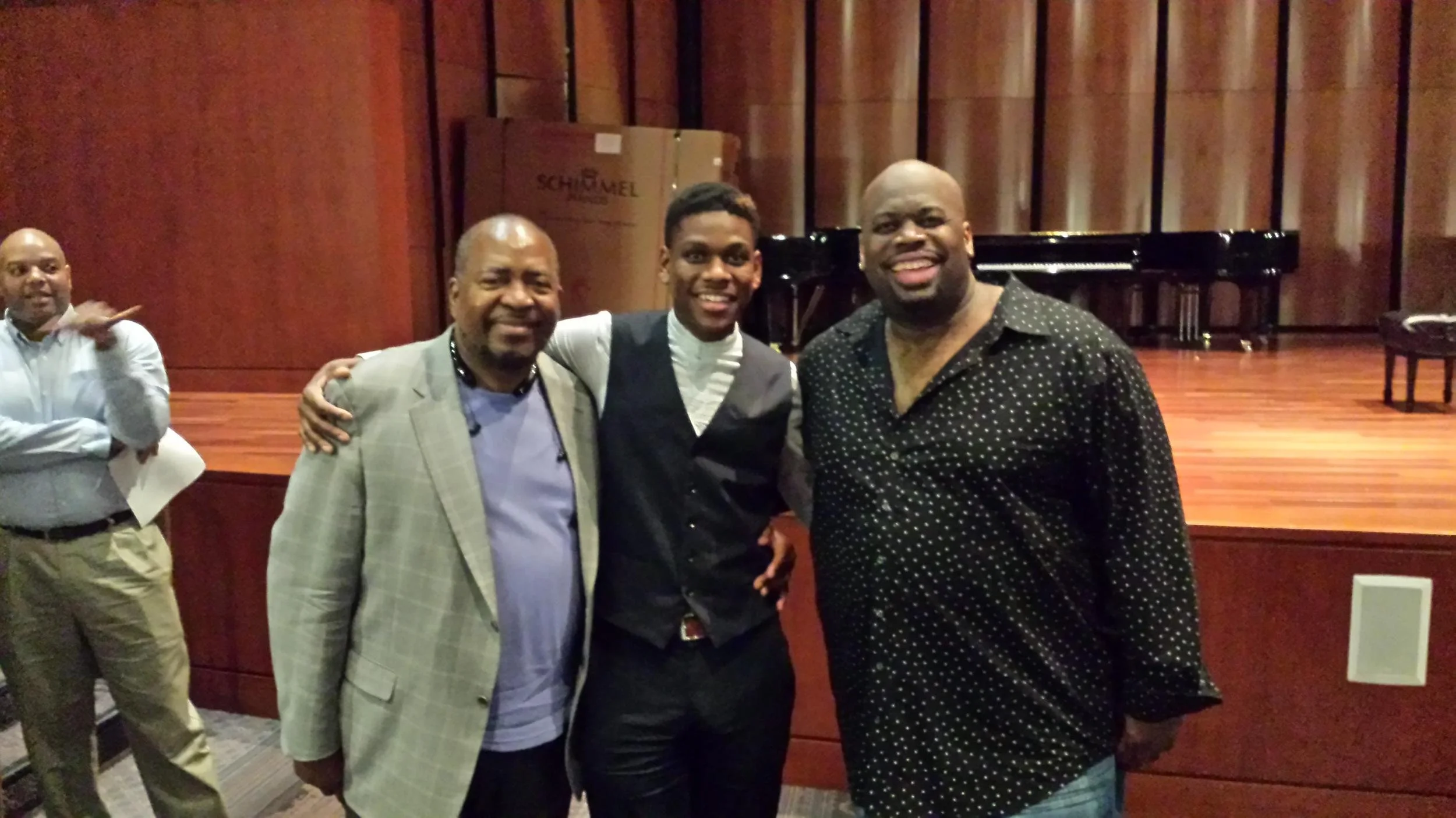 Three men standing together and smiling, with a man in the background pointing at them, in a room with a wooden stage and a piano.