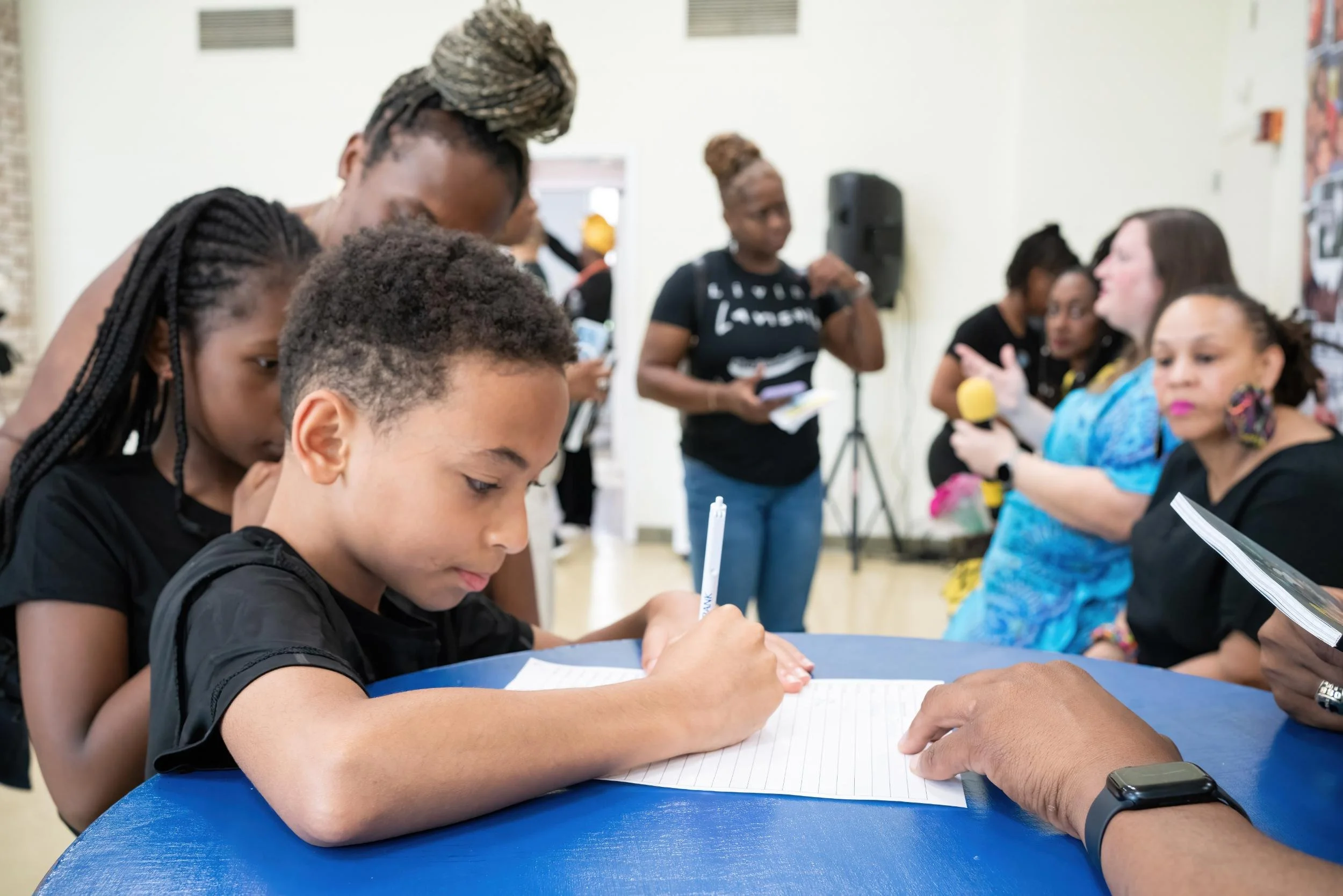 A group of children and adults gathered around a blue table in an indoor setting, with a boy in the foreground writing on a notepad, surrounded by people who appear to be participating in a workshop or community event.