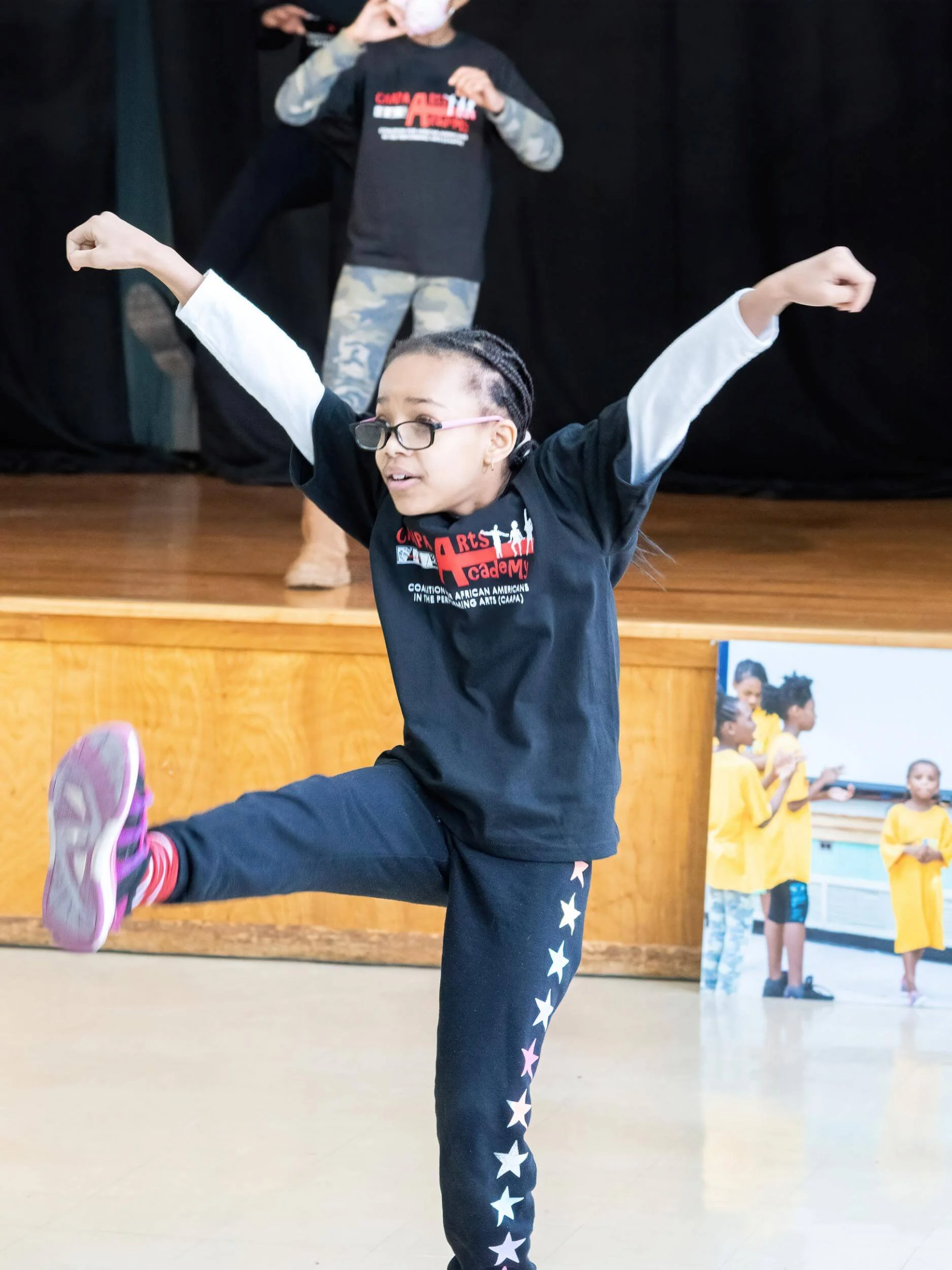 Young girl with glasses and braided hair performing a dance move on stage, raising her arms and lifting one leg, with a black background and other children in yellow shirts watching in the background.