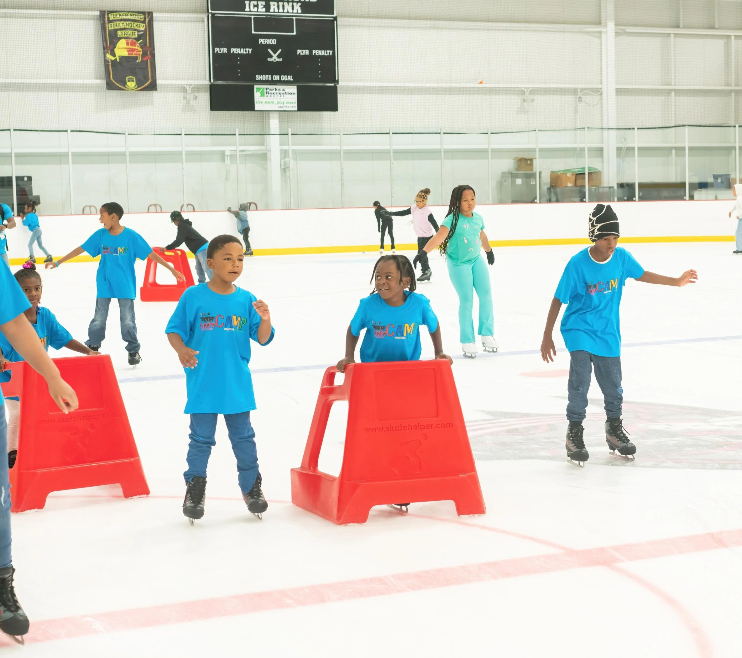 Children in blue shirts skating on an indoor ice rink, some using red skating aids, with other skaters in the background.